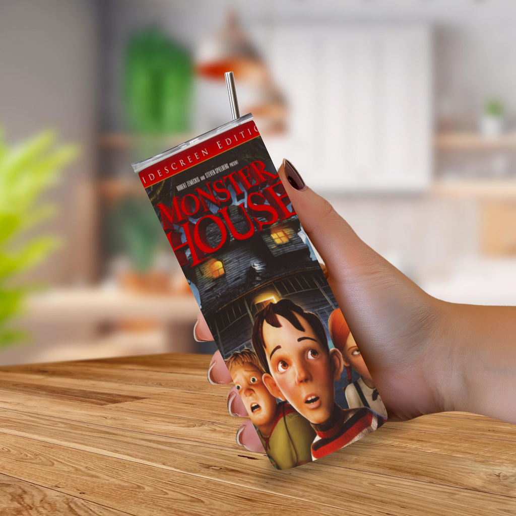 A hand holding a custom-printed skinny tumbler featuring the 'Monster House' movie artwork, with a metal straw, set against a blurred kitchen background.