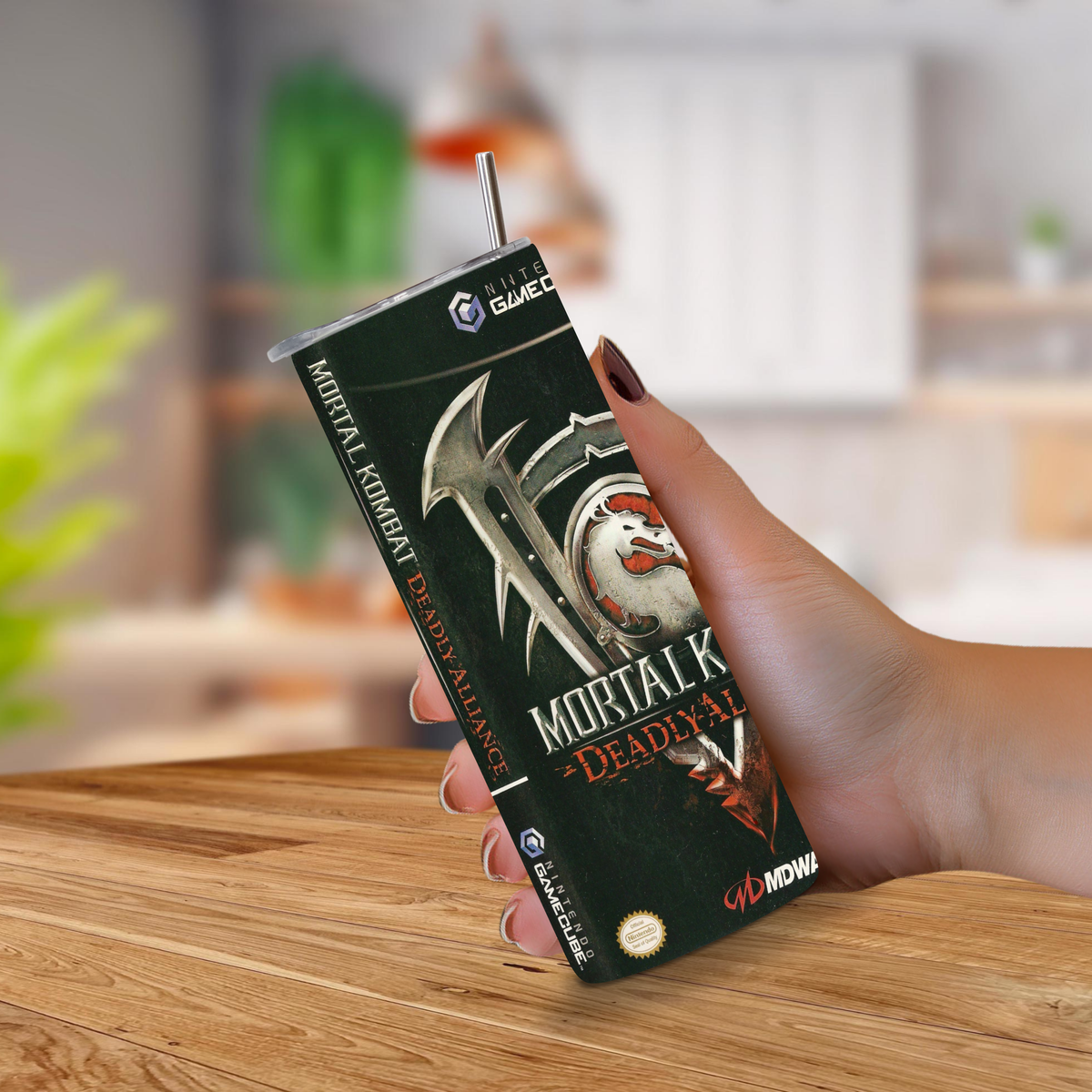 A hand holding a Mortal Kombat Deadly Alliance 20oz stainless steel tumbler in a kitchen setting with a wooden table in the foreground.
