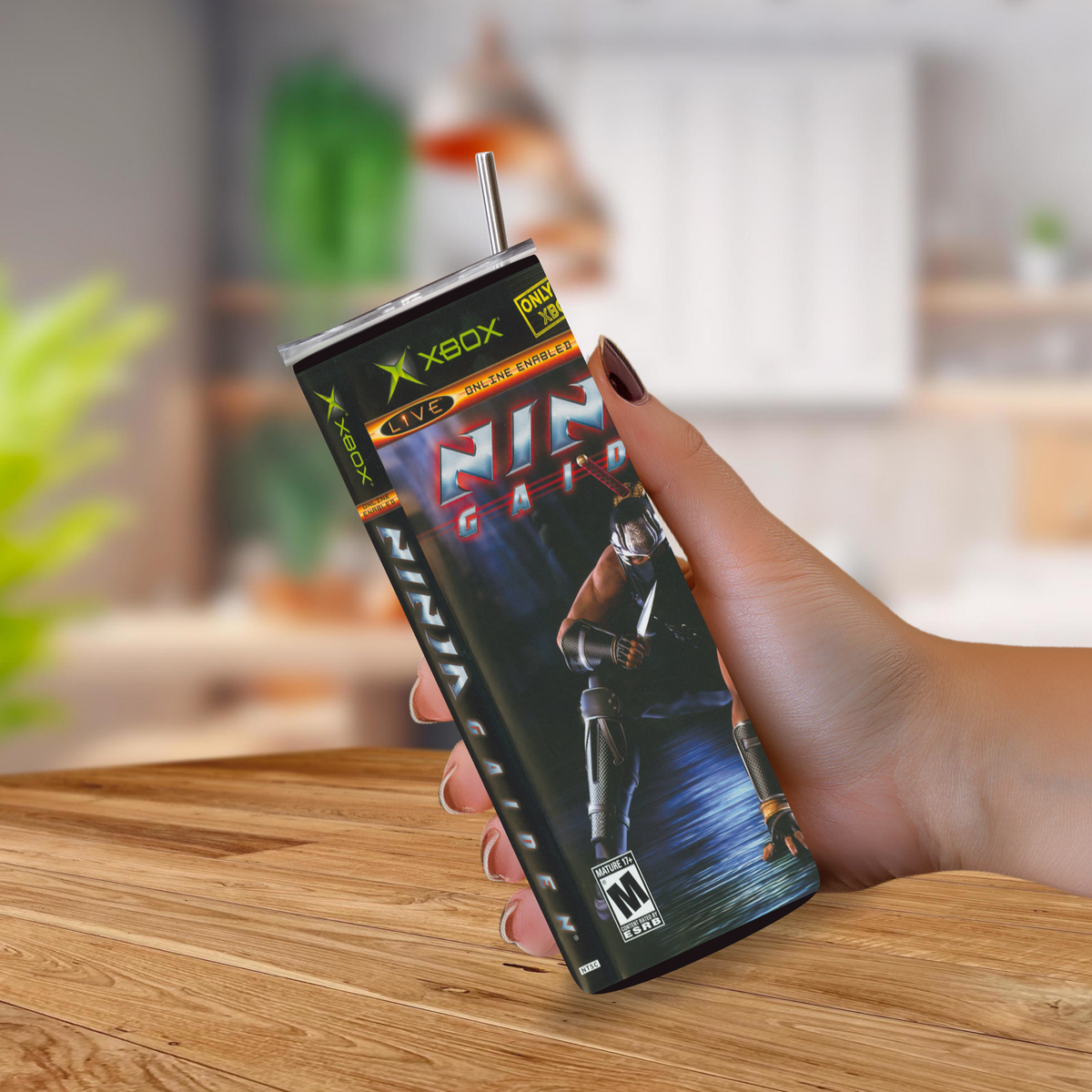 A hand holding a 20oz tumbler drinkware item that looks like an Xbox One game case "Ninja Gaiden" featuring a metallic straw, set against a blurred indoor background with a wooden table.