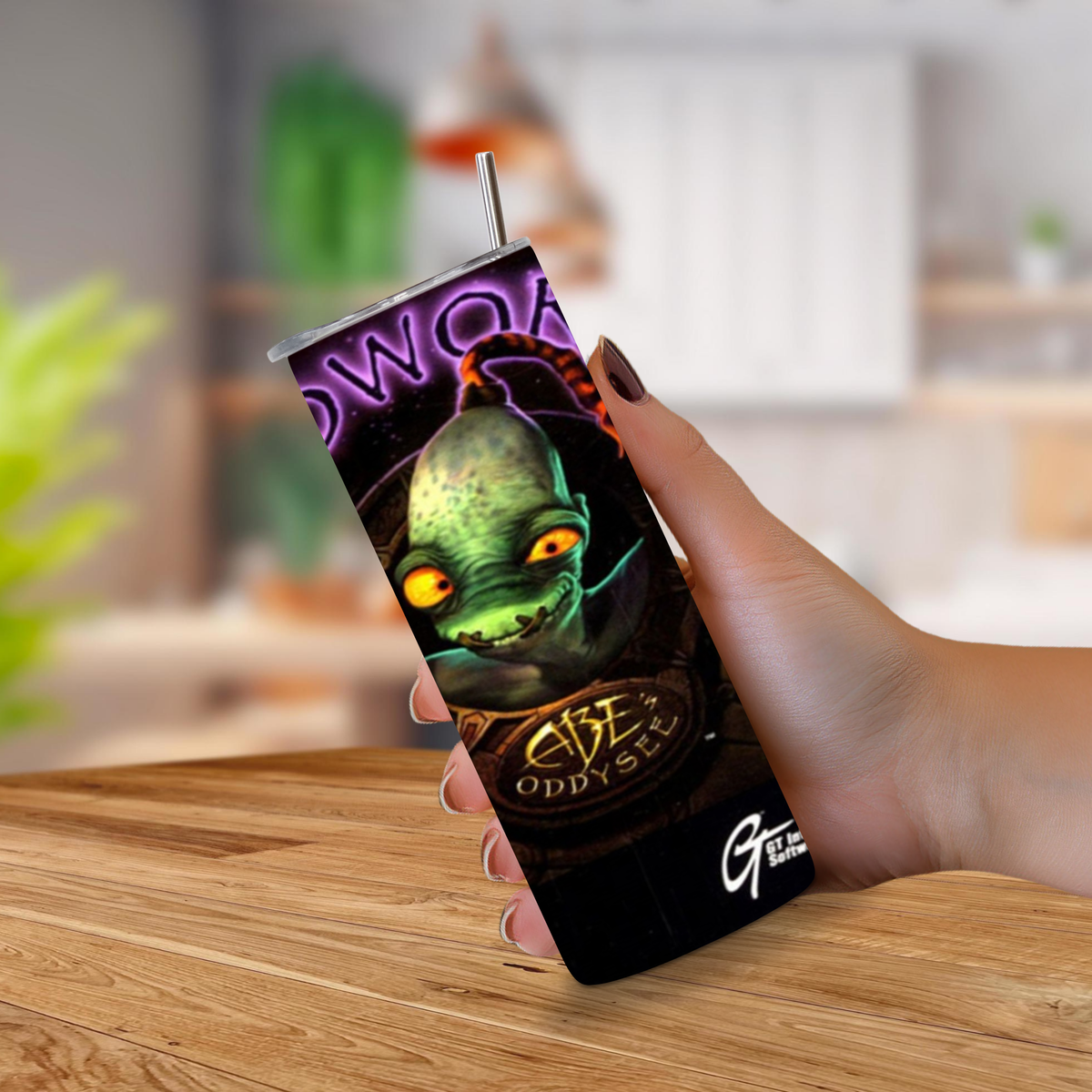 A hand holding a slim, black insulated 20oz stainless steel tumbler with a straw, featuring a colorful graphic of a green alien with glowing yellow eyes and the text 'Oddworld Abe Oddysee. The background shows a blurred indoor setting with a wooden table surface.