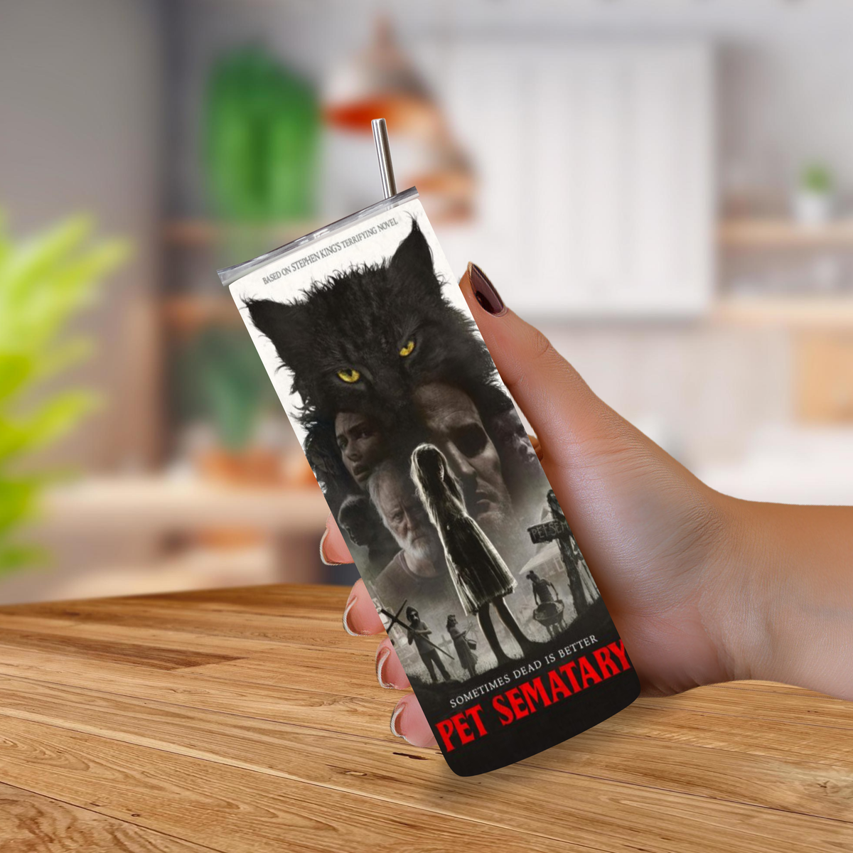 A hand holding a slim rectangular tumbler with a metal straw, printed with a black-and-white wolf and face graphic and the red text 'PET SEMATARY'. The background shows a blurred indoor room with a wooden table in the foreground.