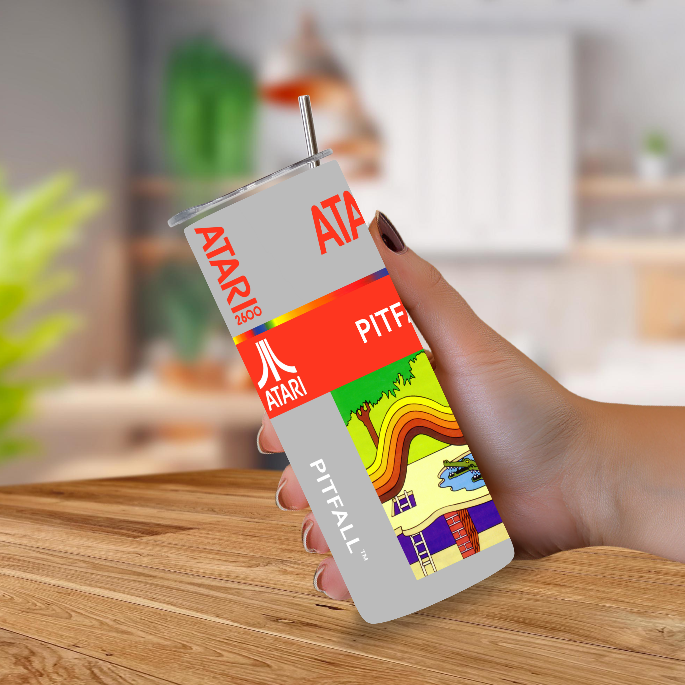 A hand holding a 20oz stainless steel tumbler with a printed Atari Pitfall design, featuring the red Atari logo and a colorful game-themed graphic, set against a blurred kitchen background.