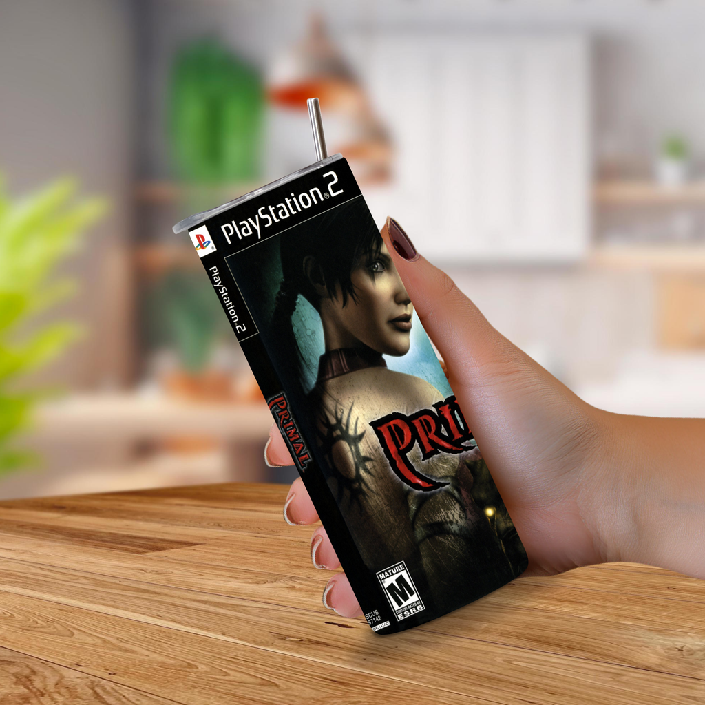A hand holding a PlayStation 2 20oz tumbler Primal design with a metal straw sticking out of the top, set against a blurred indoor background and wooden tabletop.