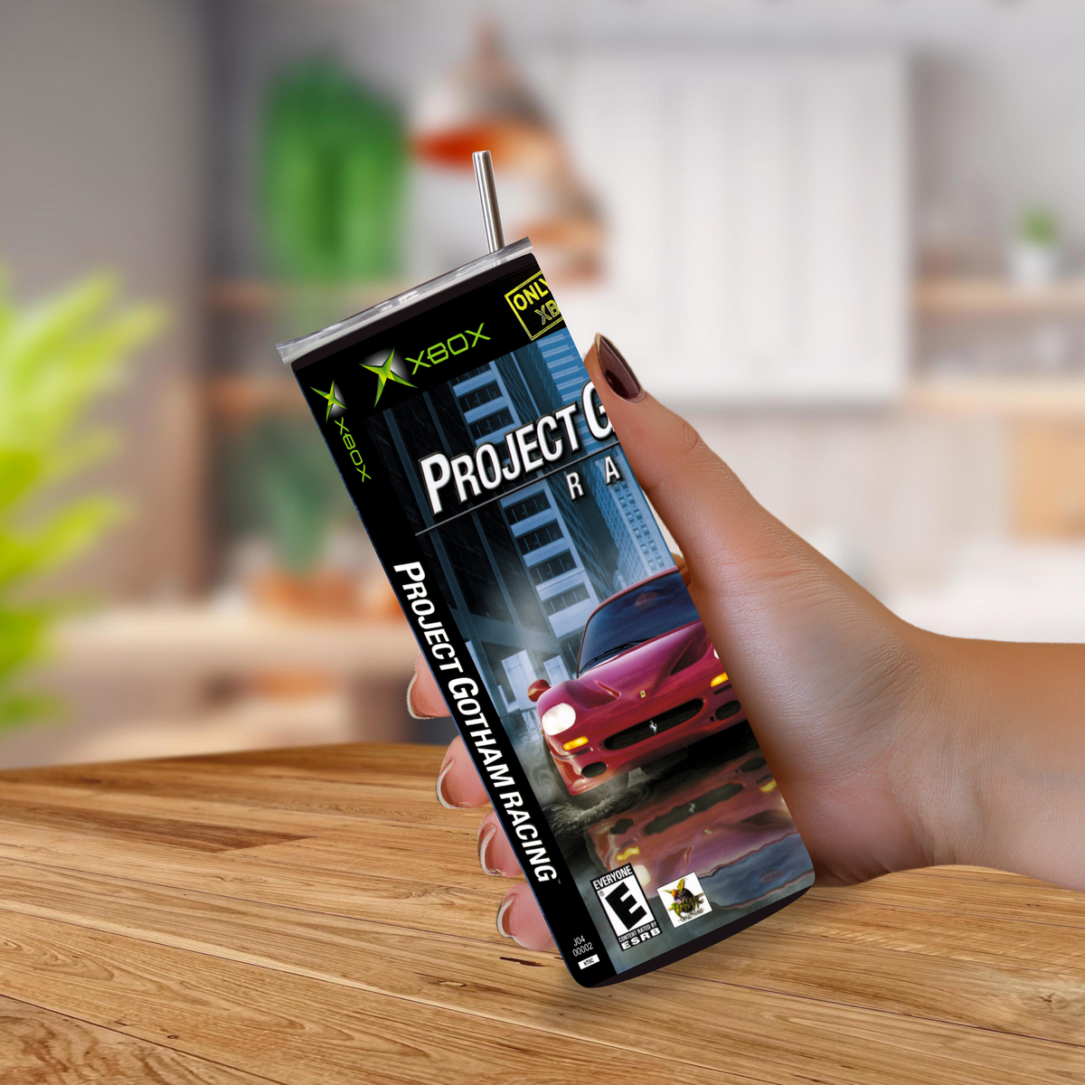 A hand holding a 20oz tumbler styled to look like the Xbox One game cover for Project Gotham Racing, with a straw inserted at the top, set against a blurred indoor background.