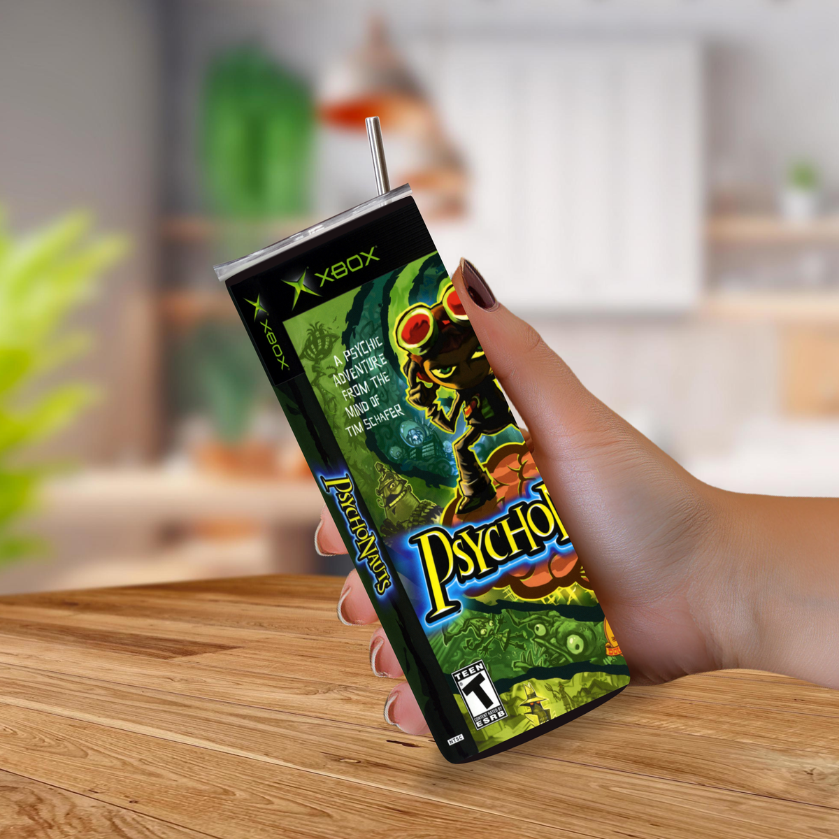 A hand holding a 20oz tumbler designed to look like the Xbox Psychonauts game, with a metal straw, set on a wooden table with a blurred kitchen background.
