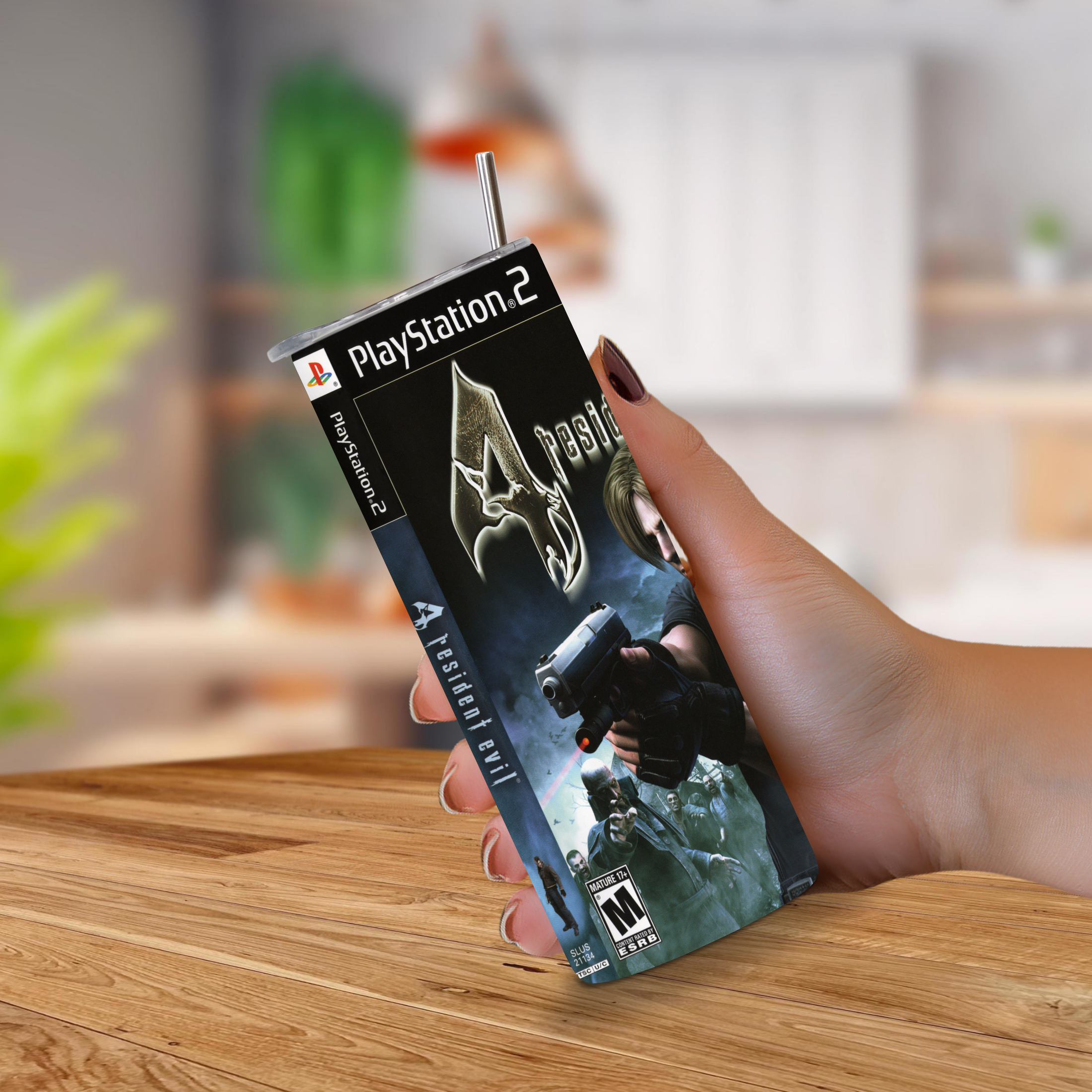 A hand holding a PlayStation 2 game of Resident Evil 4 20oz stainless steel tumbler with a metal straw extending from the top, set against a blurred indoor background and wooden tabletop.