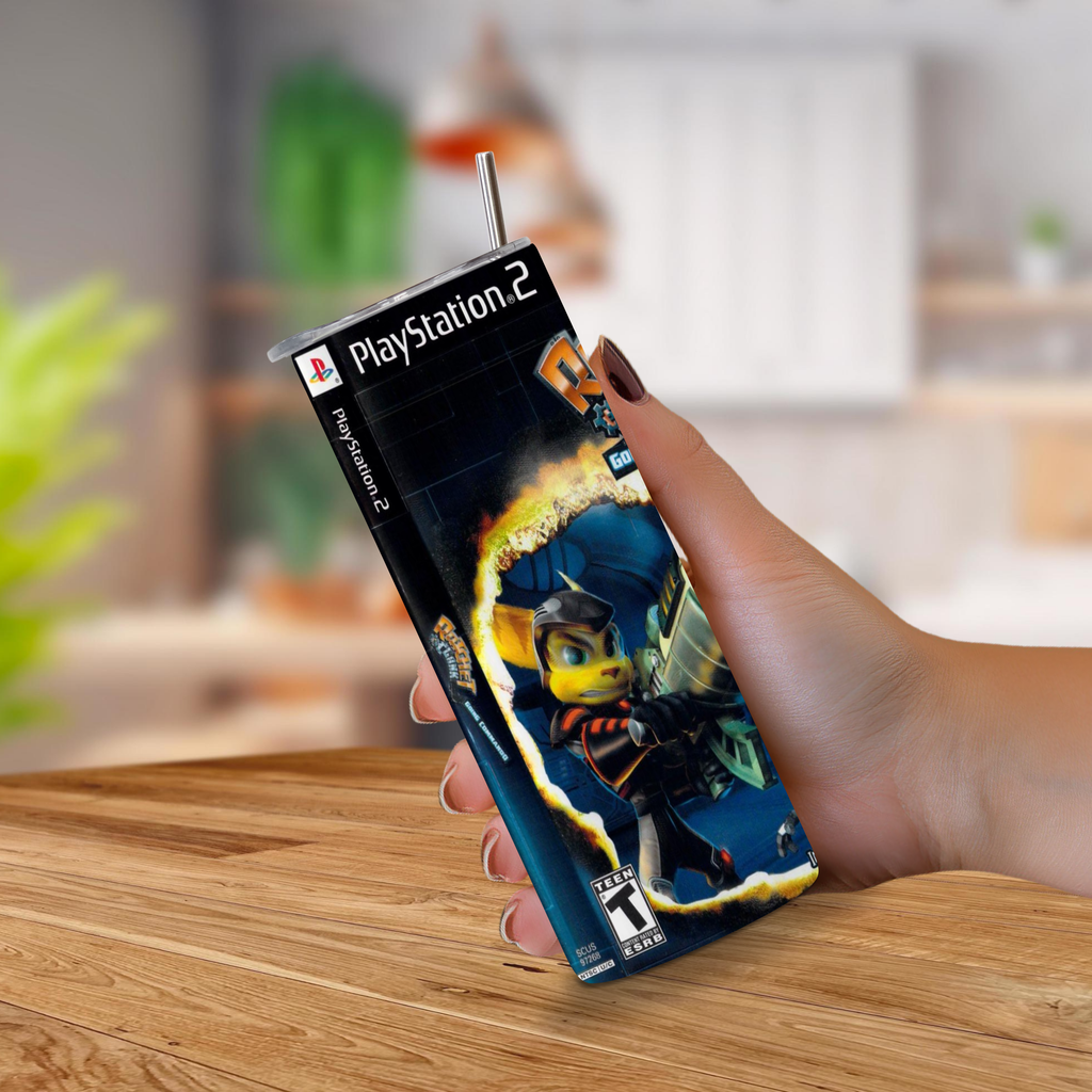 A hand holding a PlayStation 2 game cover of ratchet & clank going commando designed to look a 20oz stainless steel tumbler, with a metal straw sticking out from the top, set against a blurred indoor background.