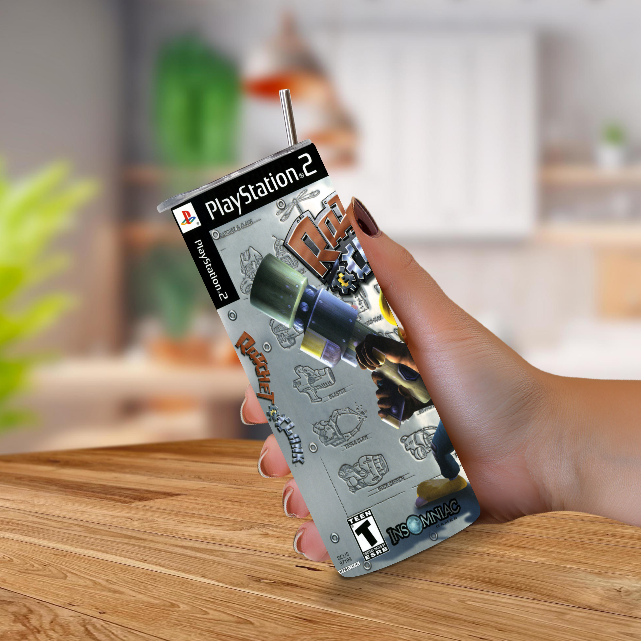 A hand holding a PlayStation 2-themed novelty 20oz stainless steel tumbler with a straw, featuring the 'Ratchet & Clank' cover art, set against a blurred indoor background.