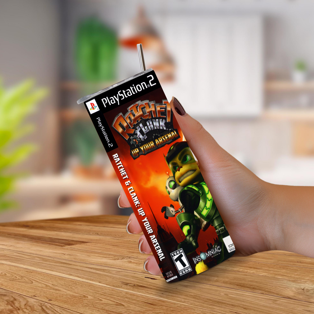 A hand holding a 20oz tumbler PlayStation 2 Ratchet & Clank: Up Your Arsenal game on a wooden table with a blurred indoor background.