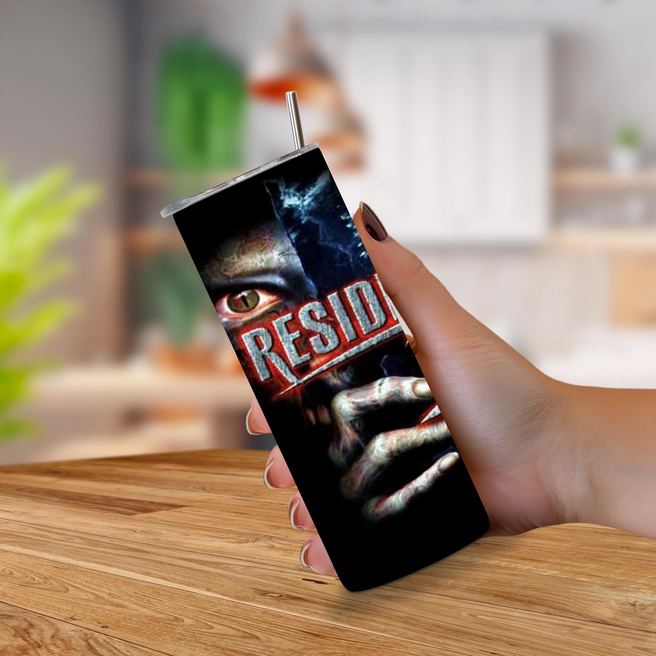 A hand holding a 20oz stainless steel tumbler with a straw, featuring a graphic print of a creepy eye and clawed hand with the word 'RESIDENT EVIL' in red, against a blurred indoor background.