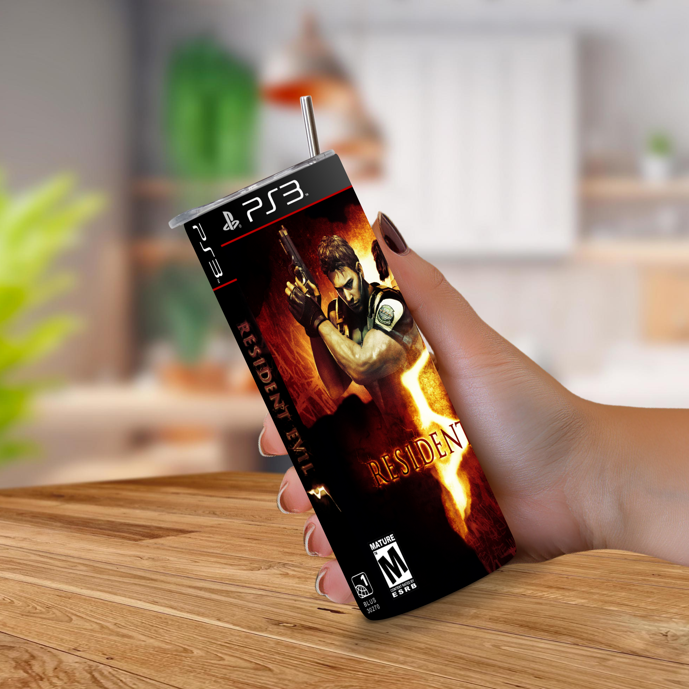 A hand holding a PS3-shaped 20oz stainless steel tumbler with a straw, printed with Resident Evil 5 artwork, set against a blurred indoor background.