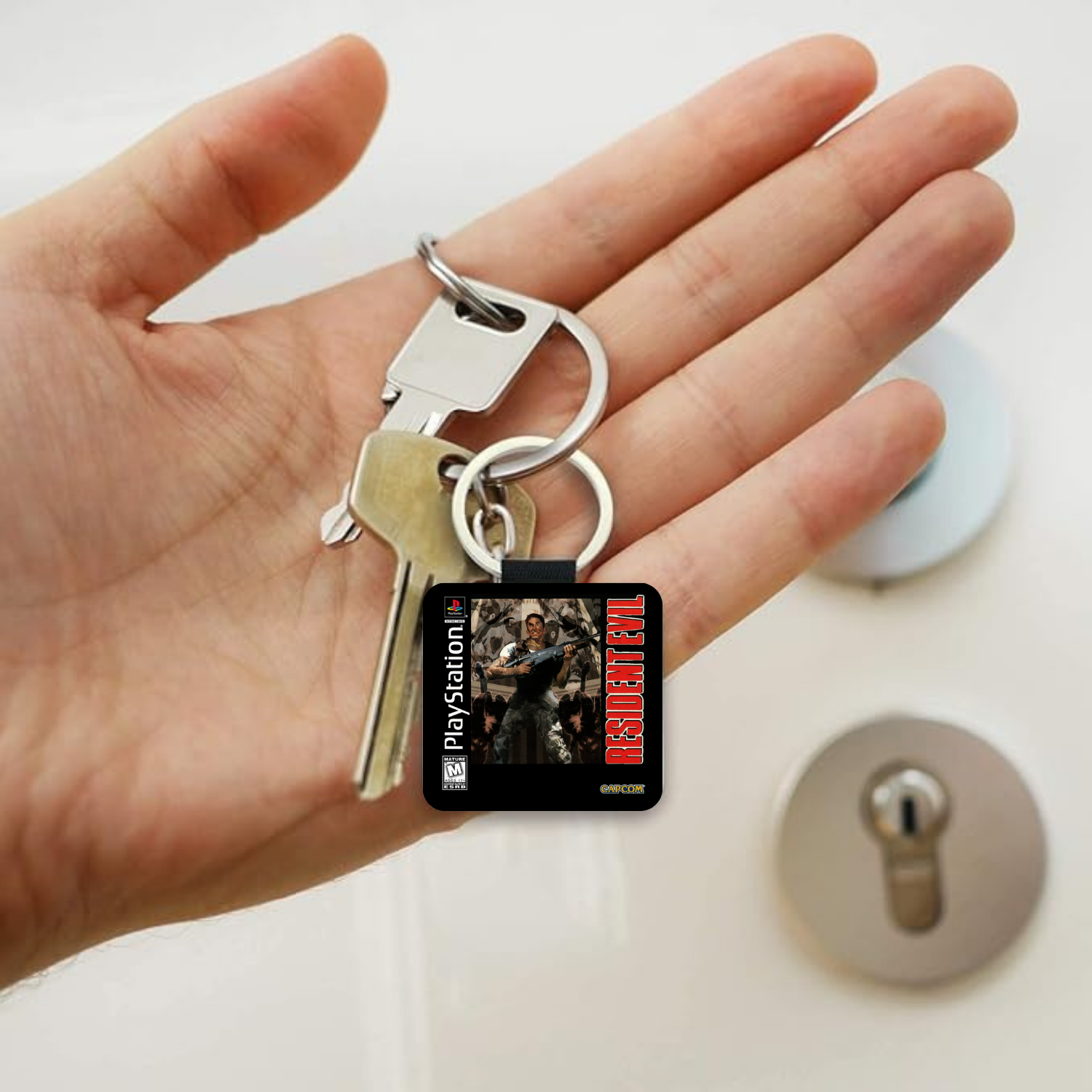 A hand holding a PlayStation 1 Resident Evil themed keychain with two keys, shown against a blurred background of a lock on a door.