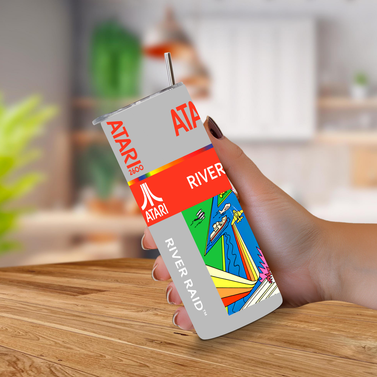 A hand holding a custom-designed Atari River Raid tumbler cup with a straw, set against a blurred kitchen background.