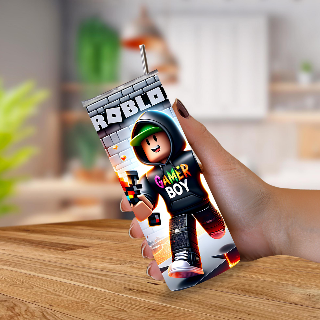 A hand holds a Roblox-branded tumbler cup with a straw, featuring a Roblox avatar graphic and the words 'Gamer Boy,' against a blurred indoor background with a wooden table.