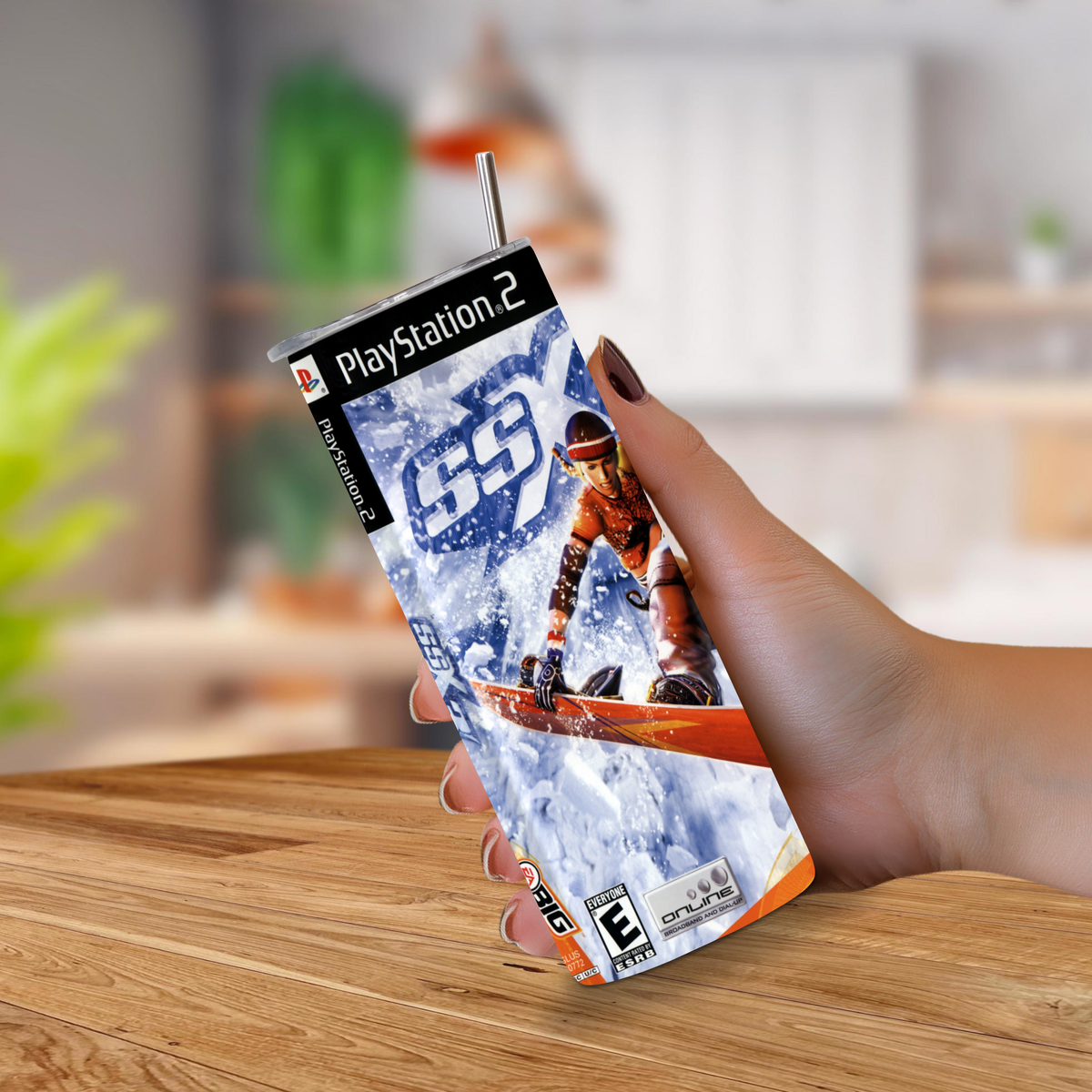 A hand holding a 20oz tumbler with PlayStation 2 game SSX 3 design with a metal straw inserted at the top, set against a blurred indoor background.