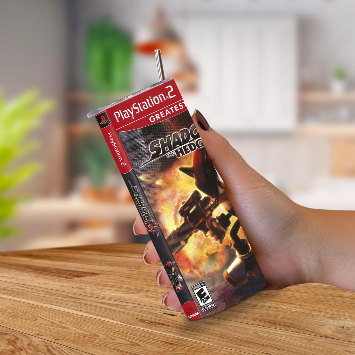 A hand holding a 20oz stainless steel tumbler PlayStation 2 game cover designed to look like the cover art for 'Shadow of the Hedgehog', with a metal straw inserted at the top, set against a blurred kitchen background.