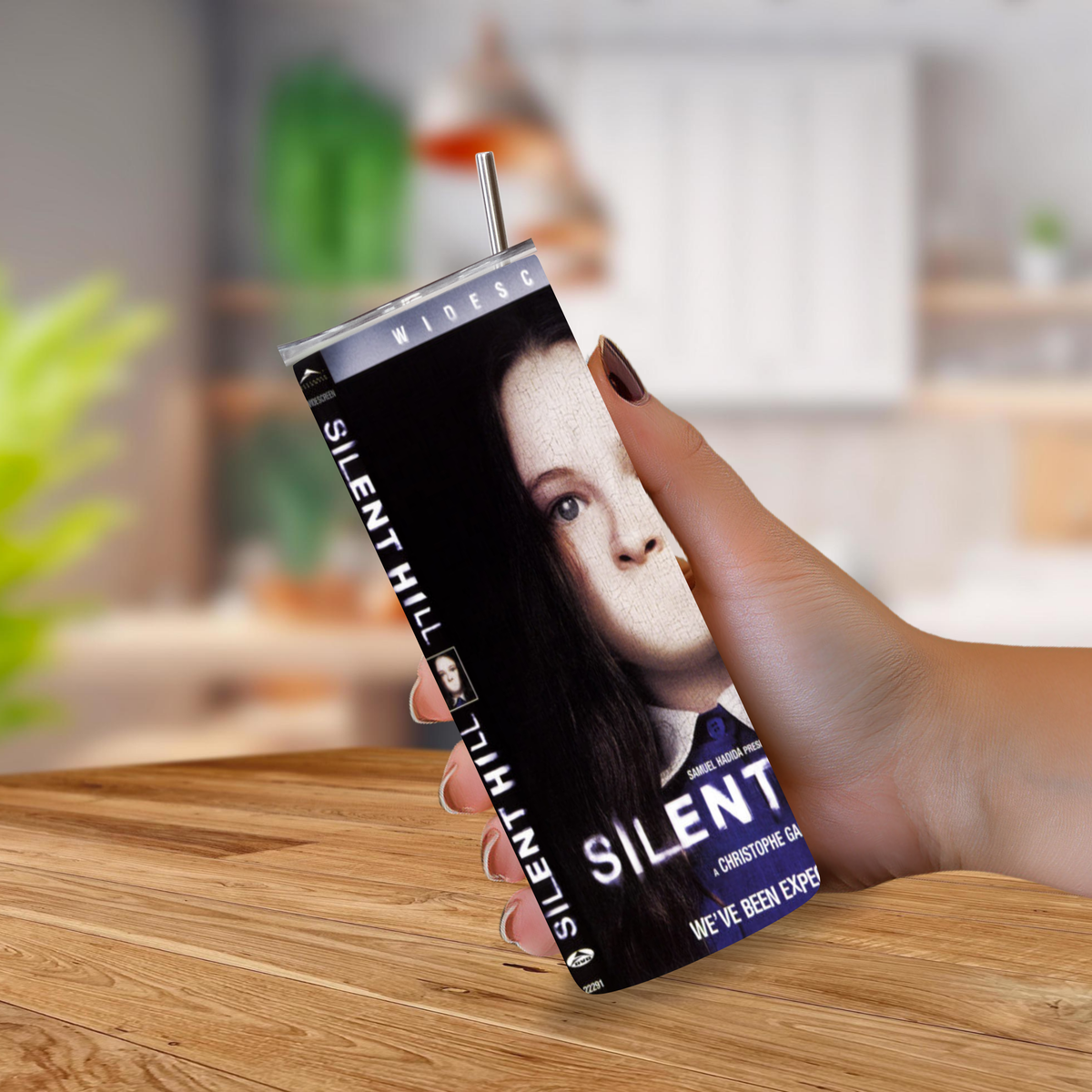 A hand holding a tall, slim 20oz tumbler with a straw, printed with the 'Silent Hill' movie design, against a blurred kitchen background.