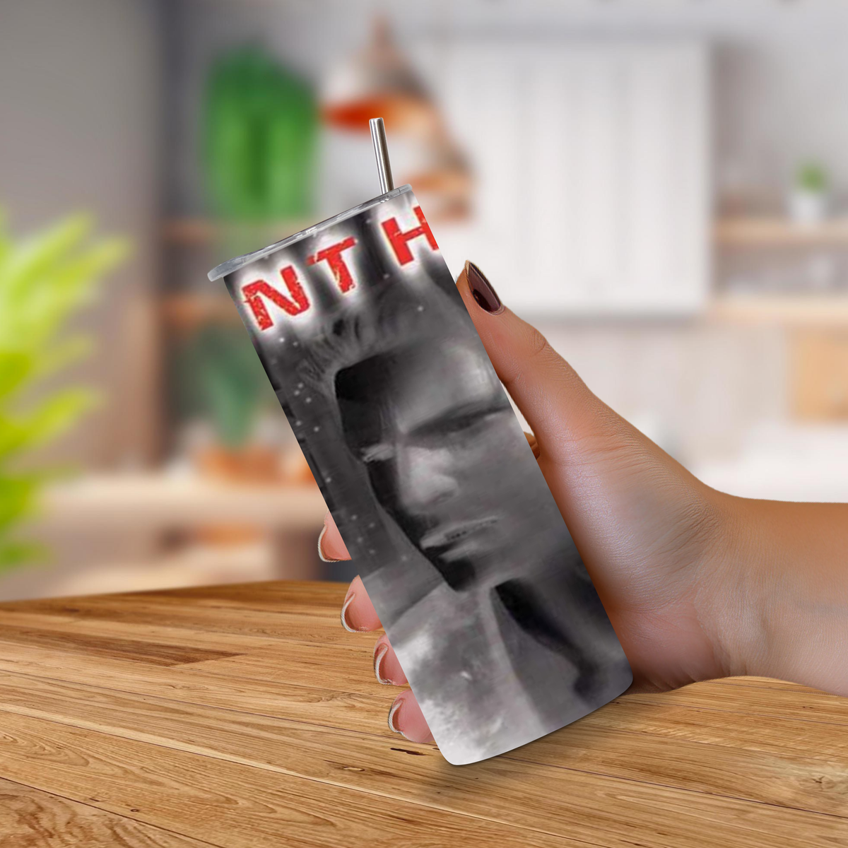 A hand holding a 20oz stainless steel tumbler with a printed black and white portrait design and red Silent Hill against a blurred kitchen background.
