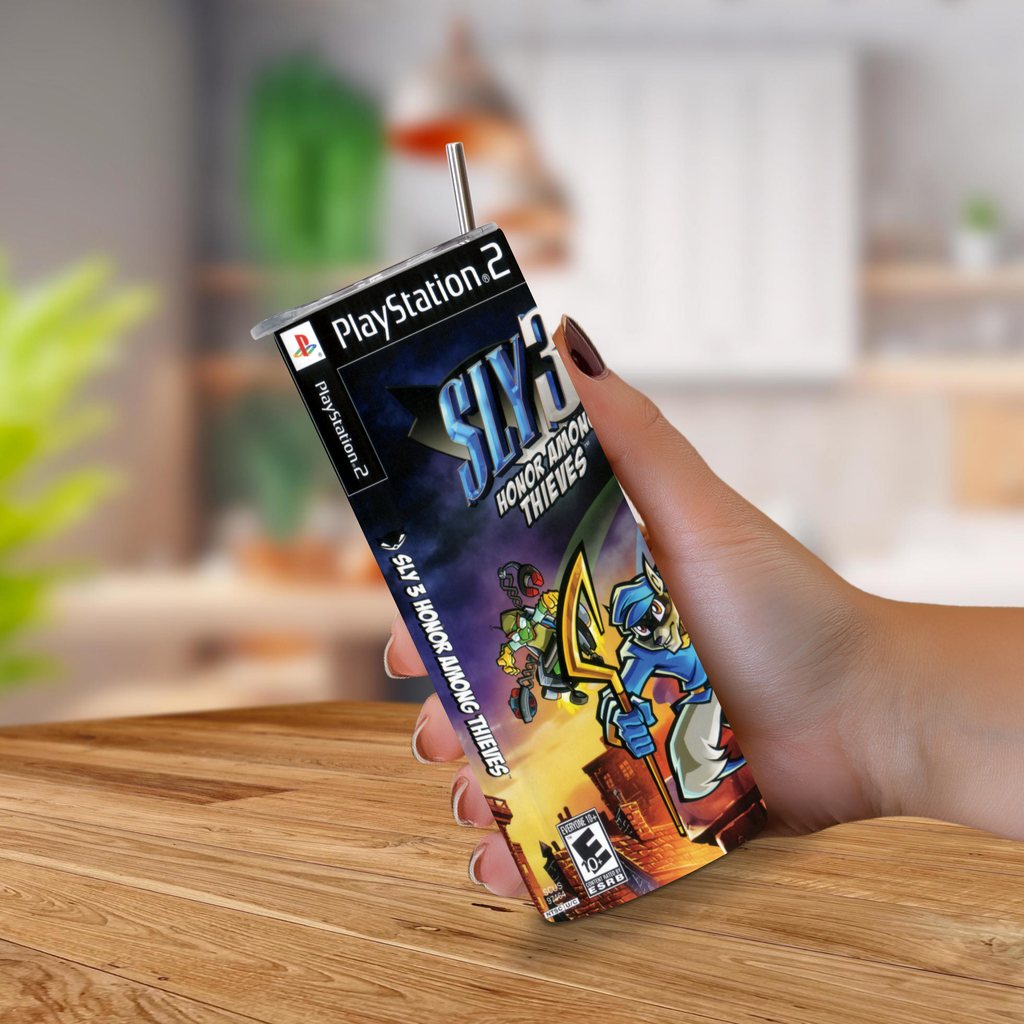 A hand holding a PlayStation 2 game cover shaped like a 20oz stainless steel tumbler featuring Sly 3 Honor Among Thieves, with a straw inserted at the top, against a blurred indoor background and wooden tabletop