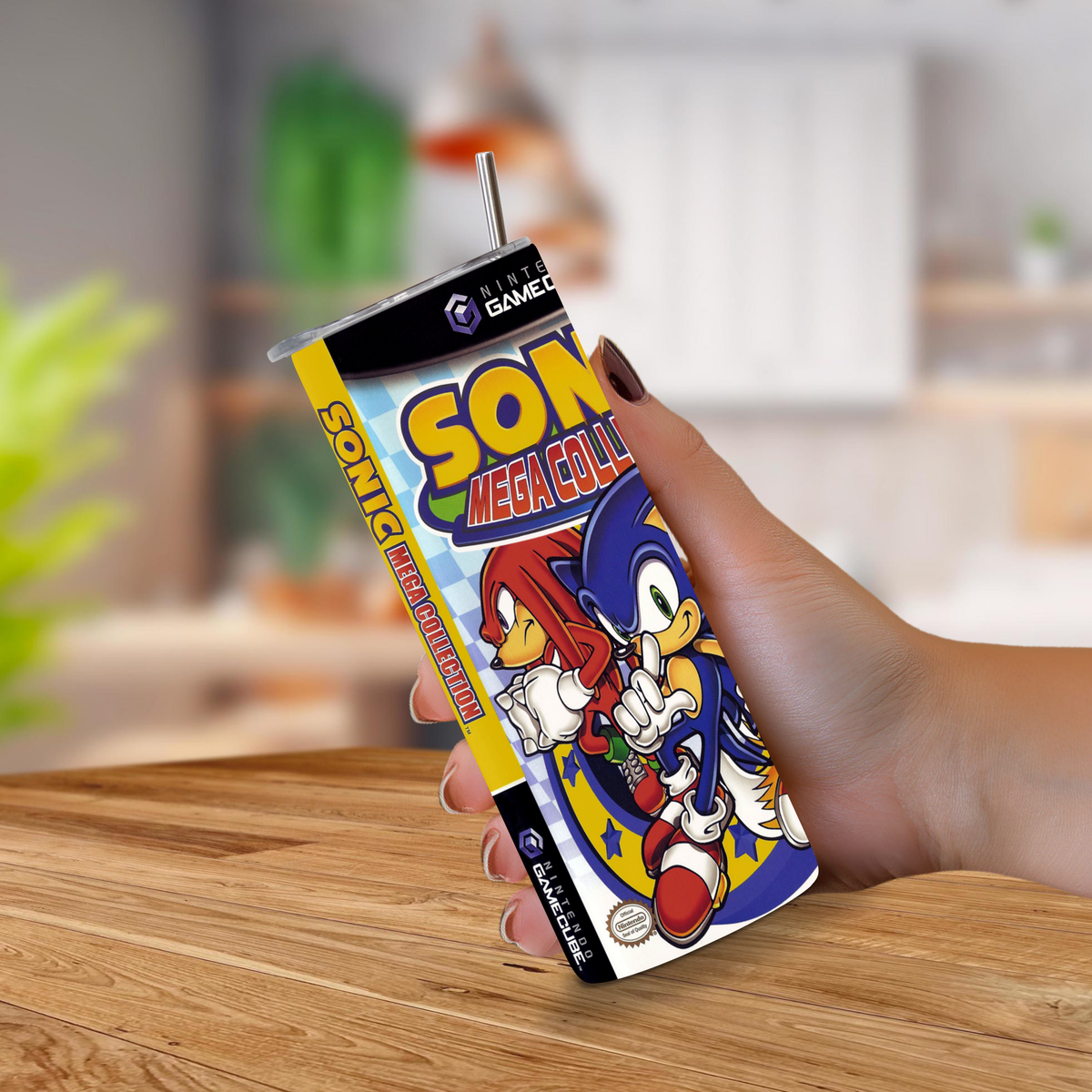 A hand holding a 20 stainless steel tumbler with a printed Sonic Mega Collection sleeve design, featuring Sonic and Knuckles, with a metal straw, set against a blurred kitchen background.