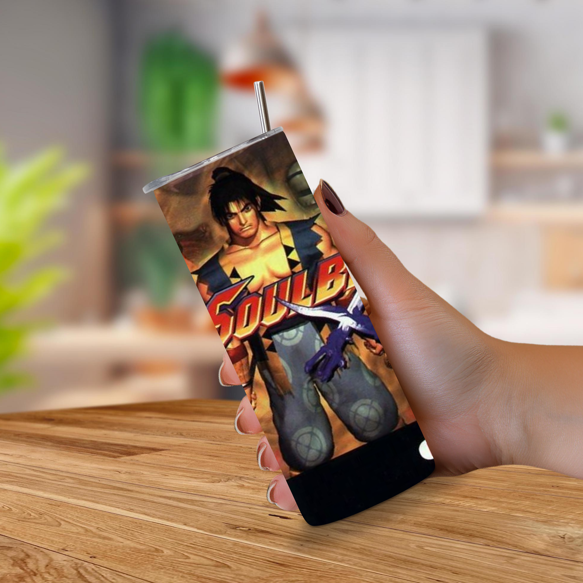 A hand holding a 20oz stainless steel tumbler with a printed Soul Blade design, featuring a metal straw, set against a blurred indoor background with a wooden table.