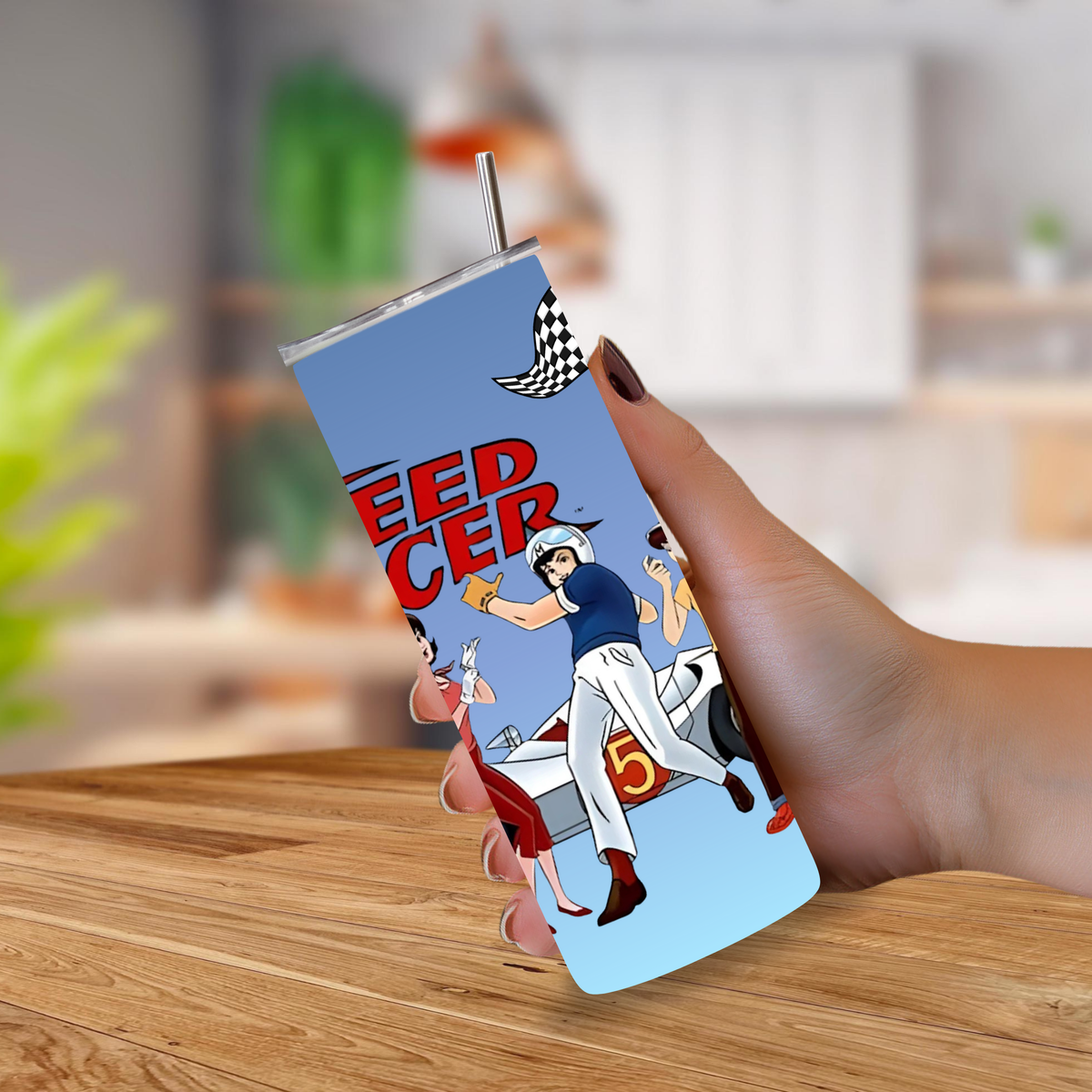 A hand holding a 20oz tumbler with a metal straw, featuring a retro-style racing illustration with the text 'Speed Racer' on a light blue background, set against a blurred kitchen backdrop.