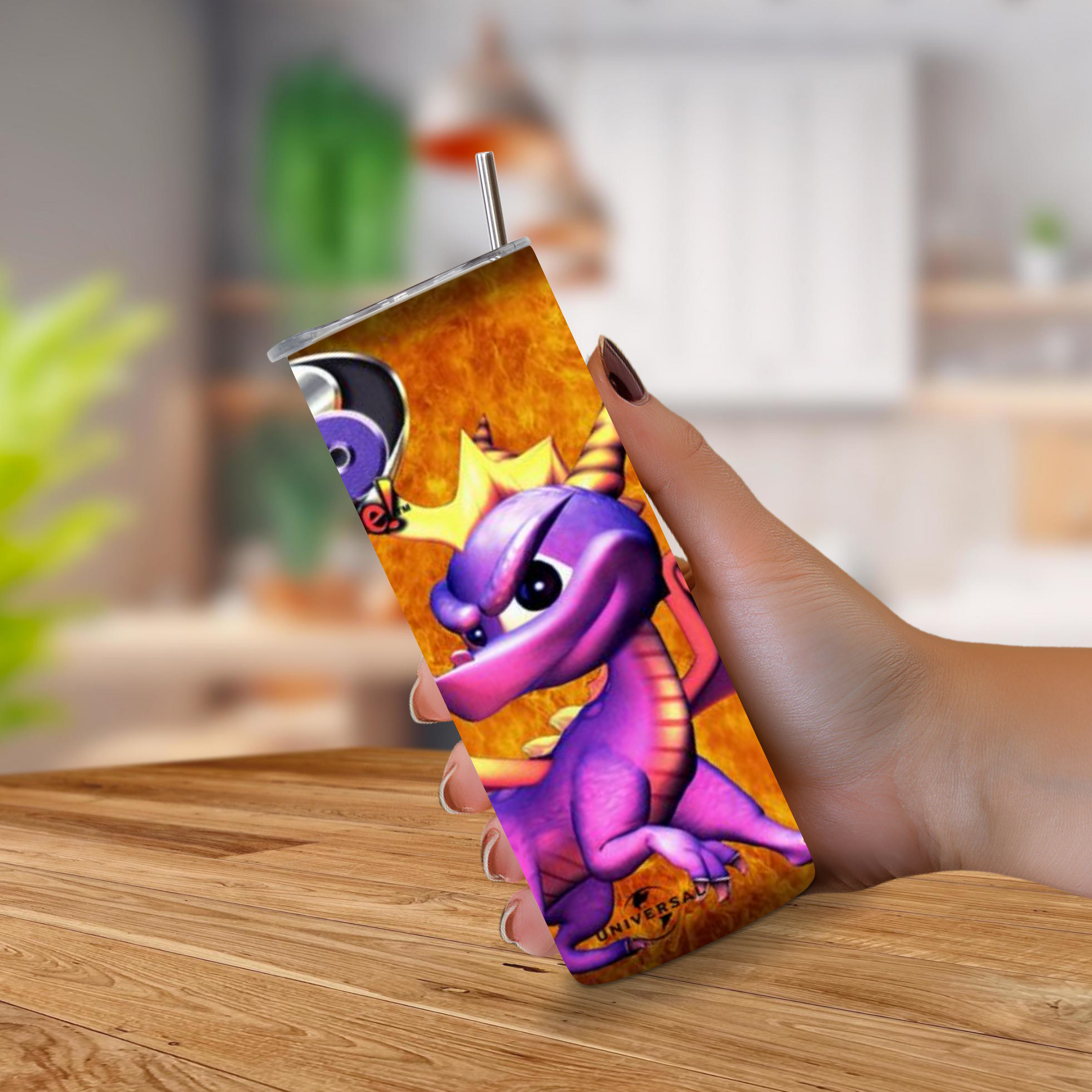 A hand holding a 20oz slim stainless steel tumbler with a printed wraparound design of Spyro 2 Ripto Rage on an orange background, with a metal straw, set against a blurred kitchen backdrop.
