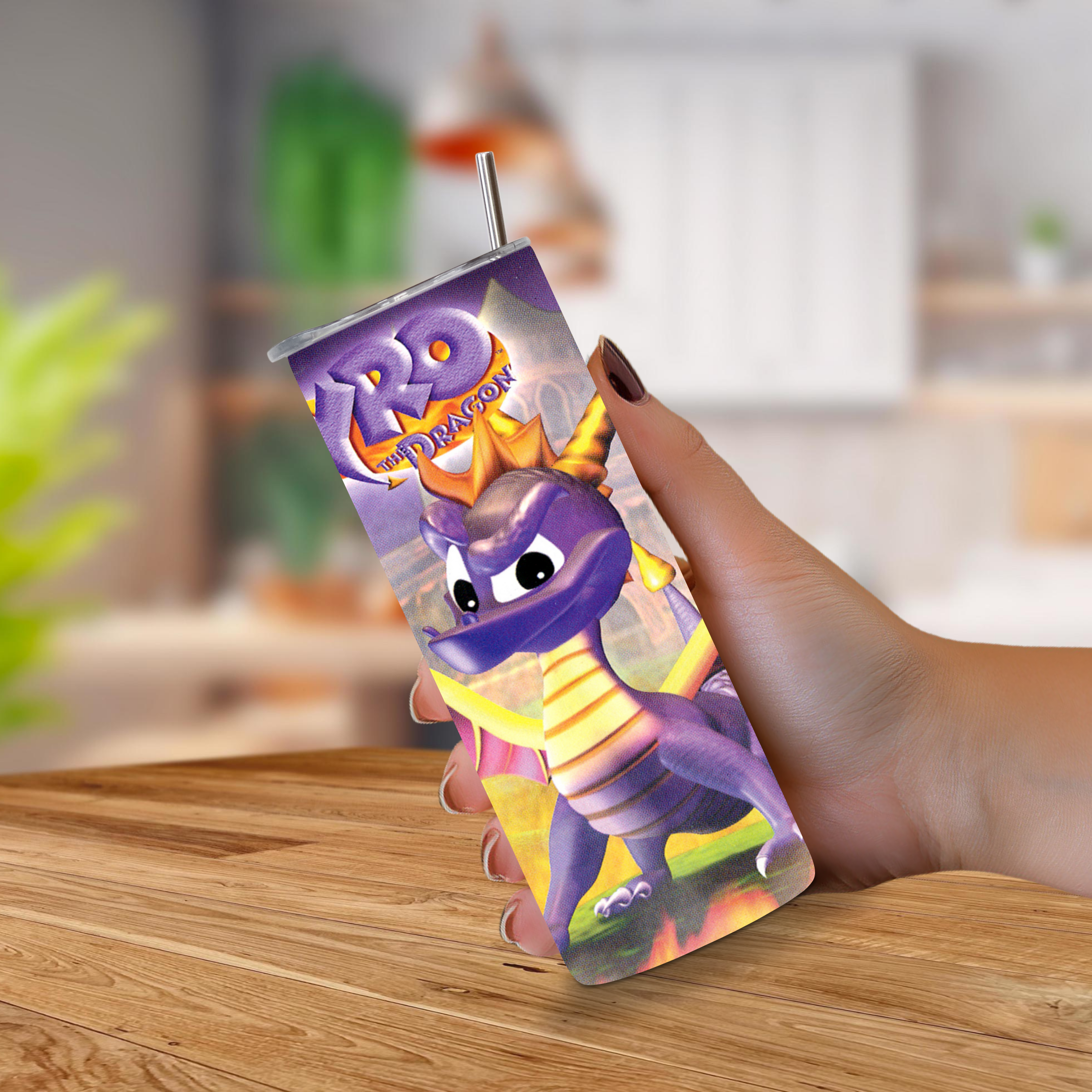 A hand holding a 20oz stainless steel tumbler with a printed Spyro the Dragon design and a metal straw, set against a blurred indoor background with a wooden table.
