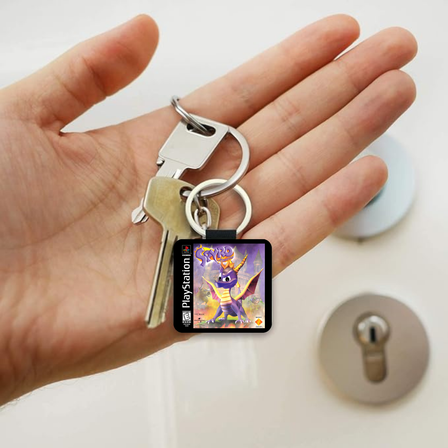 A hand holding a silver keyring with a small square PlayStation-branded charm featuring a colorful Spyro the Dragon character illustration, with a lock on a white door in the background.