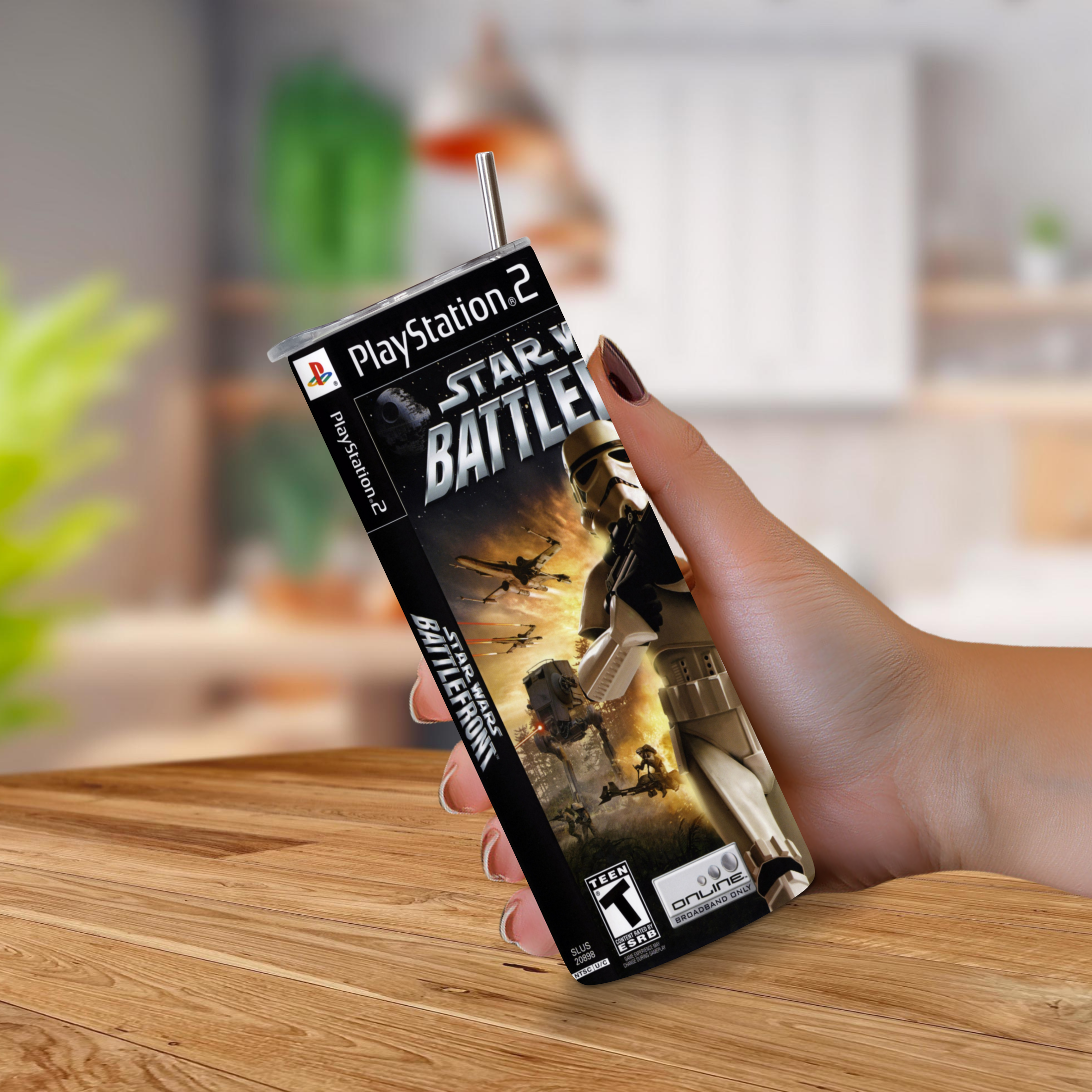 A hand holding a 20oz tumbler with PlayStation 2 Star Wars Battlefront design, with a straw inserted at the top, against a blurred indoor background.