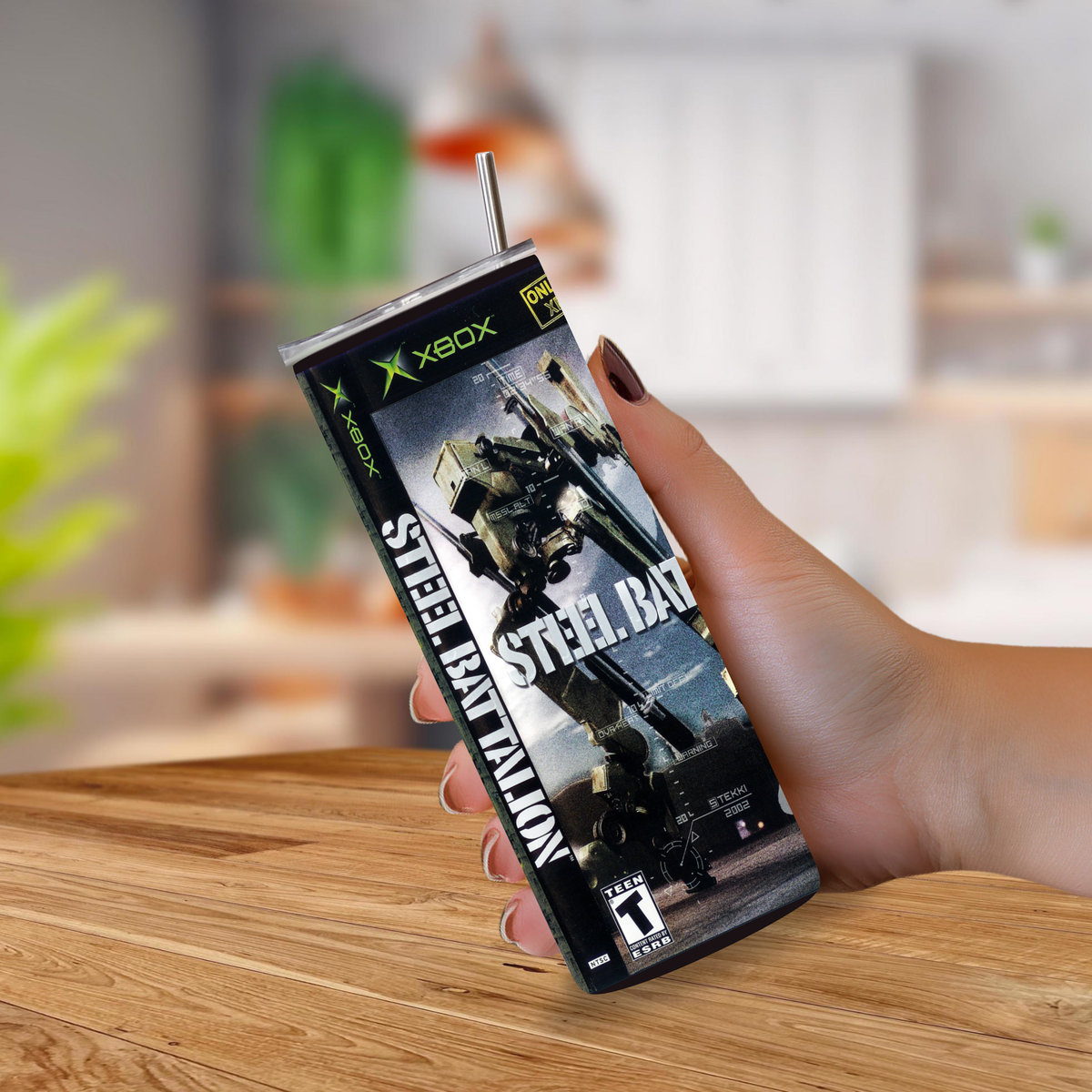 A hand holding a Steel Battalion Xbox gaming tumbler cup with a straw, set against a blurred indoor background with a wooden table.