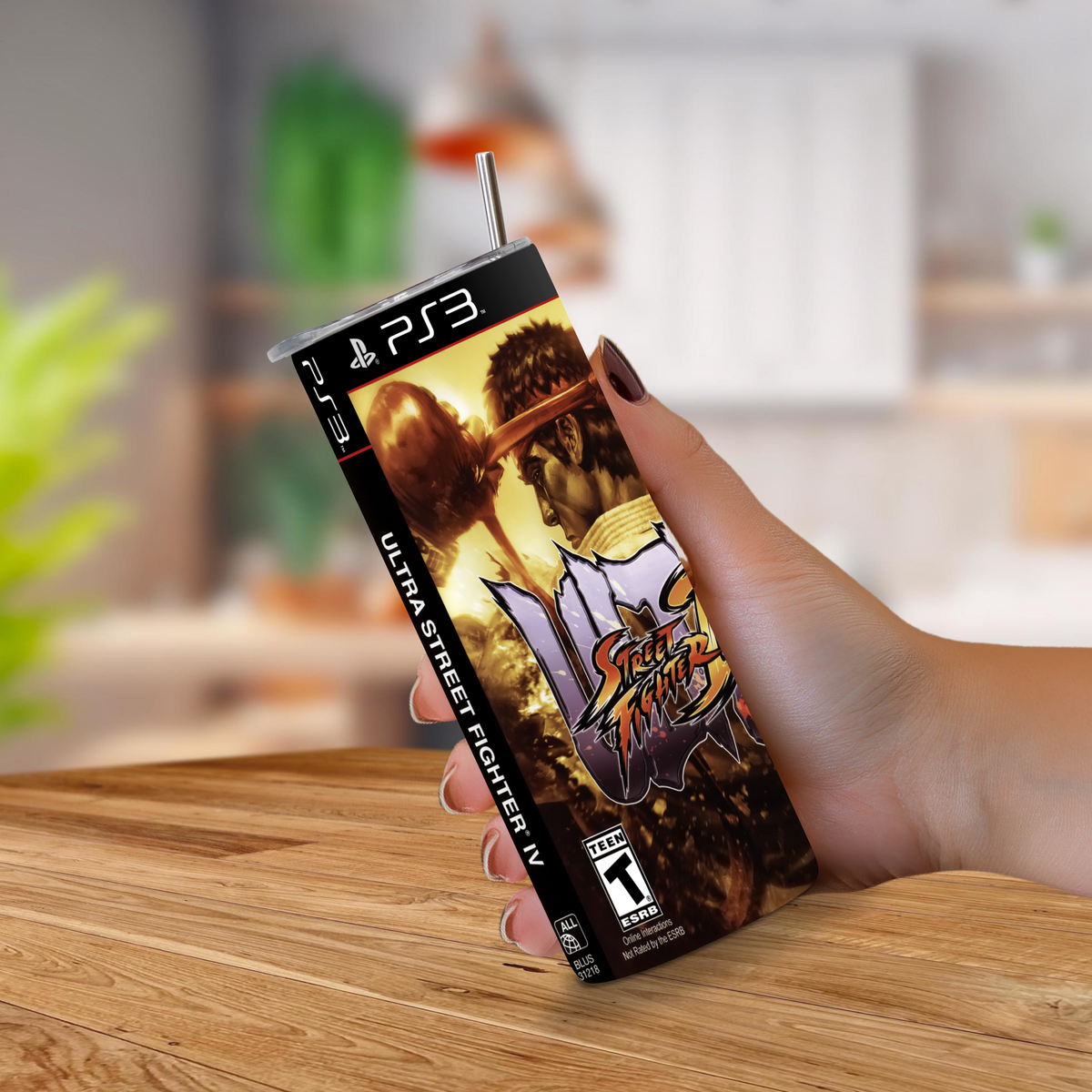A hand holding a PS3-shaped 20oz stainless steel tumbler with a straw, featuring Ultra Street Fighter IV artwork, set against a blurred indoor background with a wooden table.