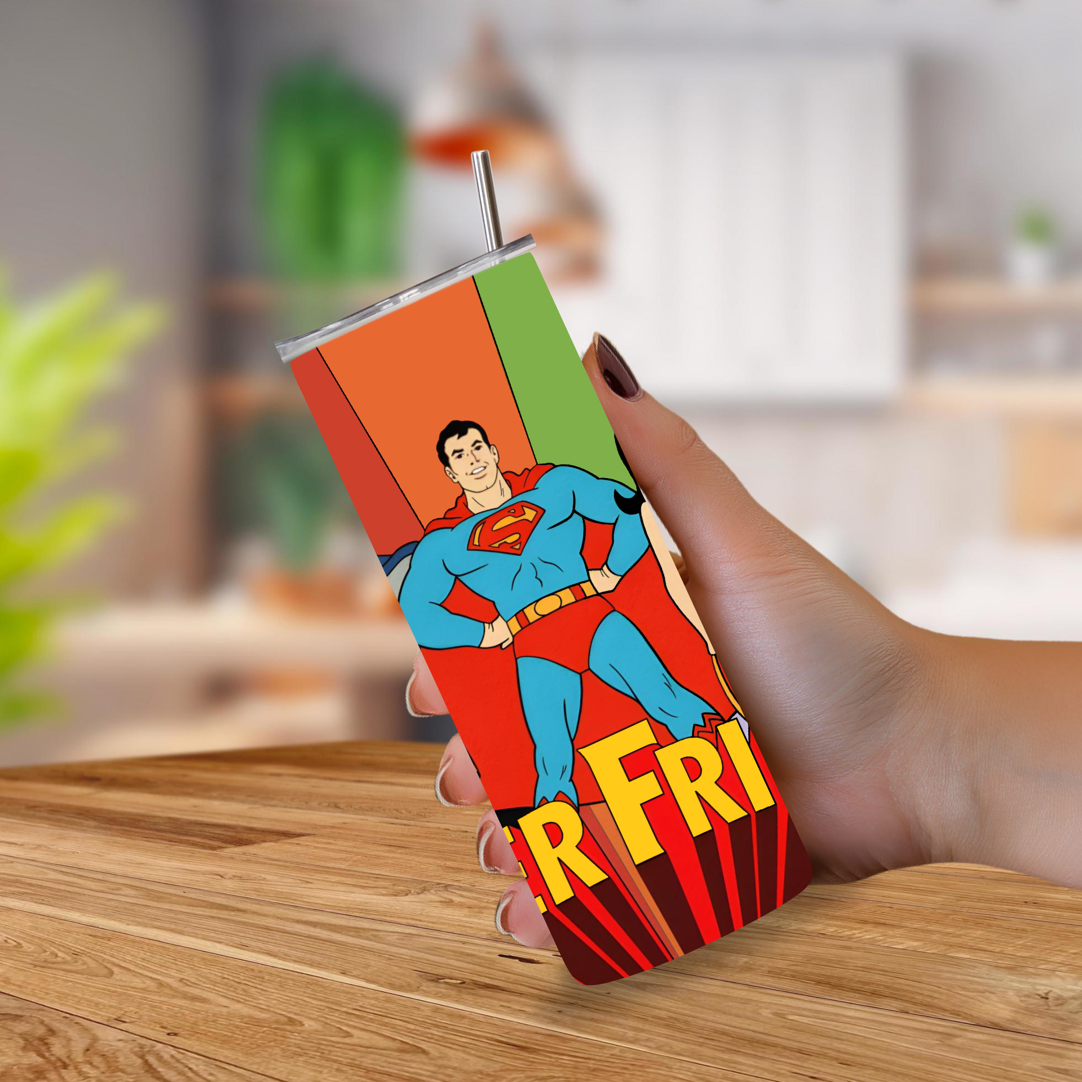 A hand holding a slim stainless-steel tumbler with a Super Friends-themed wrap design, featuring a metal straw, set against a blurred kitchen background.