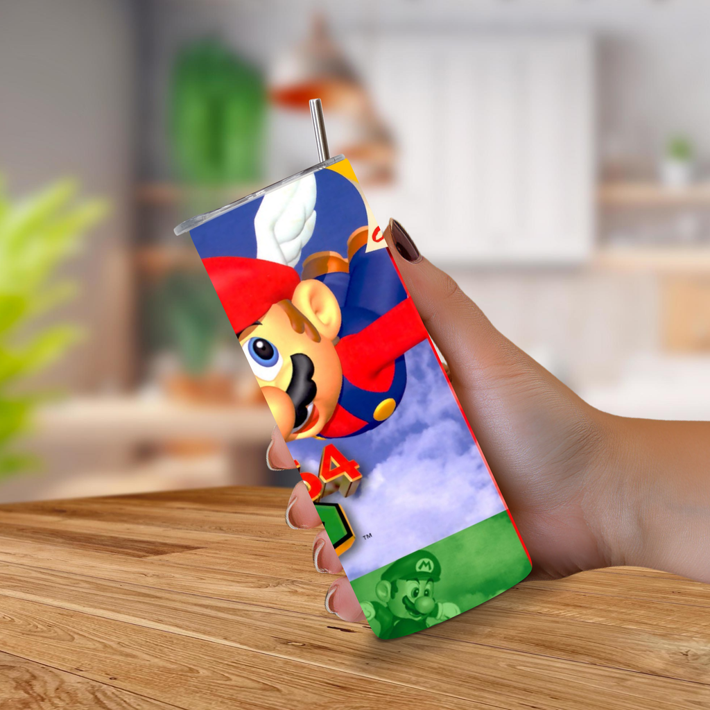 A hand holding a 20oz stainless steel tumbler featuring Super Mario 64 print, with a metal straw extending upward, set against a blurred indoor background and wooden tabletop.
