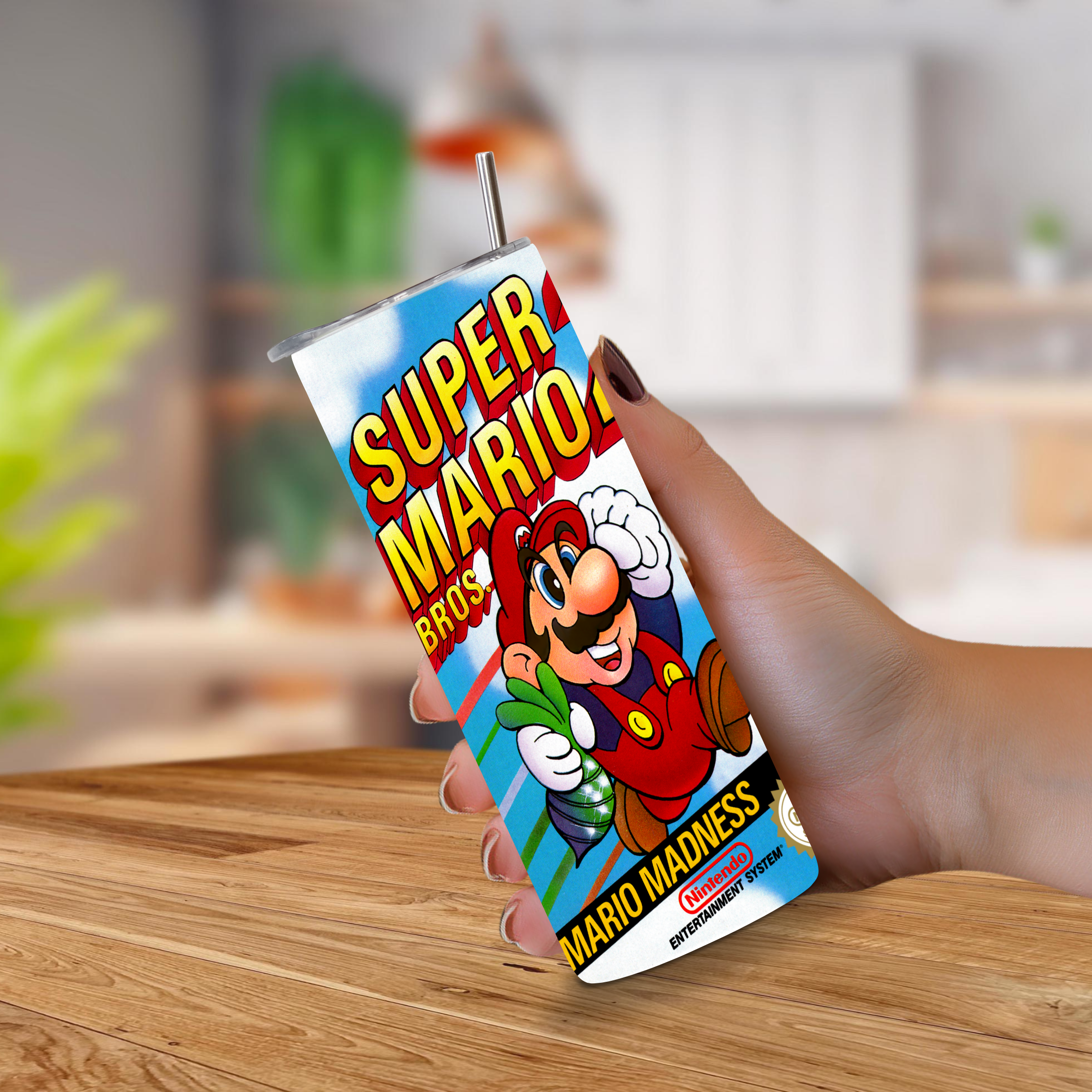 A hand holding a tall, slim 20oz tumbler with a straw featuring a colorful NES Super Mario Bros. 2 graphic print, set against a blurred indoor background with a wooden table.
