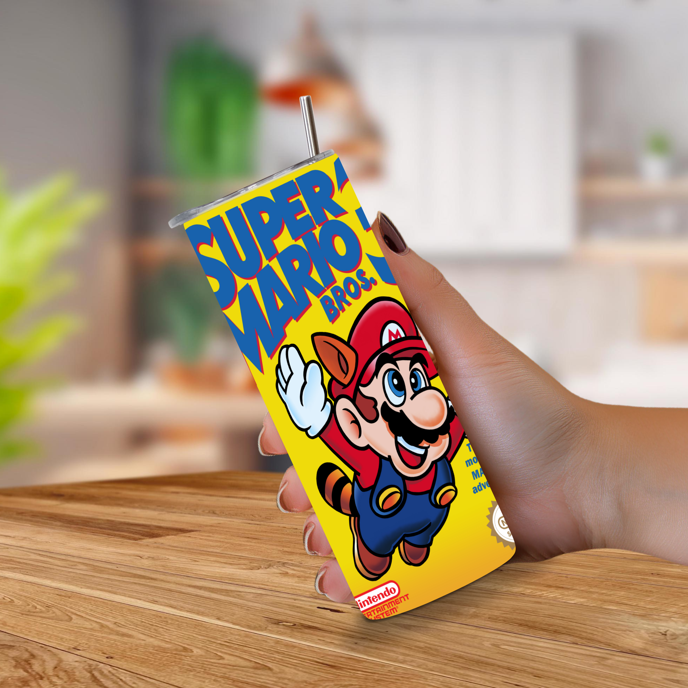 A hand holding a yellow Super Mario Bros. 3 NES designed tumbler with a straw, featuring a colorful graphic of Mario on the front, set against a blurred kitchen background.