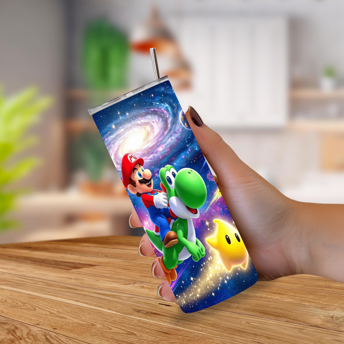 A hand holding a slim tumbler with a metal straw, printed with a colorful Super Mario and Yoshi space-themed graphic, against a blurred indoor background.