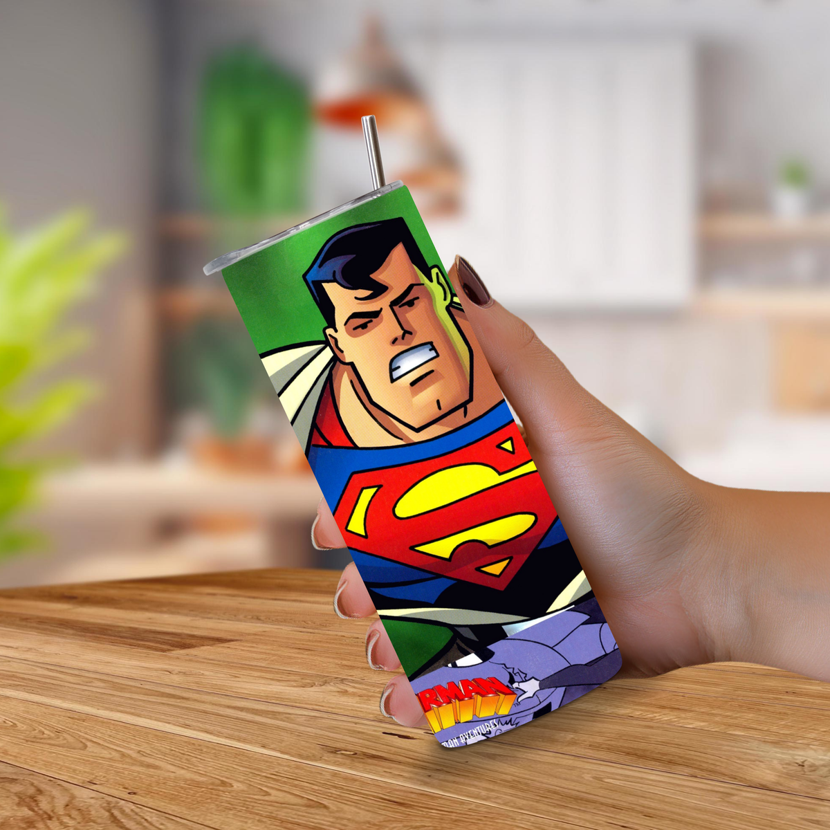 A hand holding a 20oz tumbler with a Superman N64 video game print, featuring a metal straw, set against a blurred kitchen background.