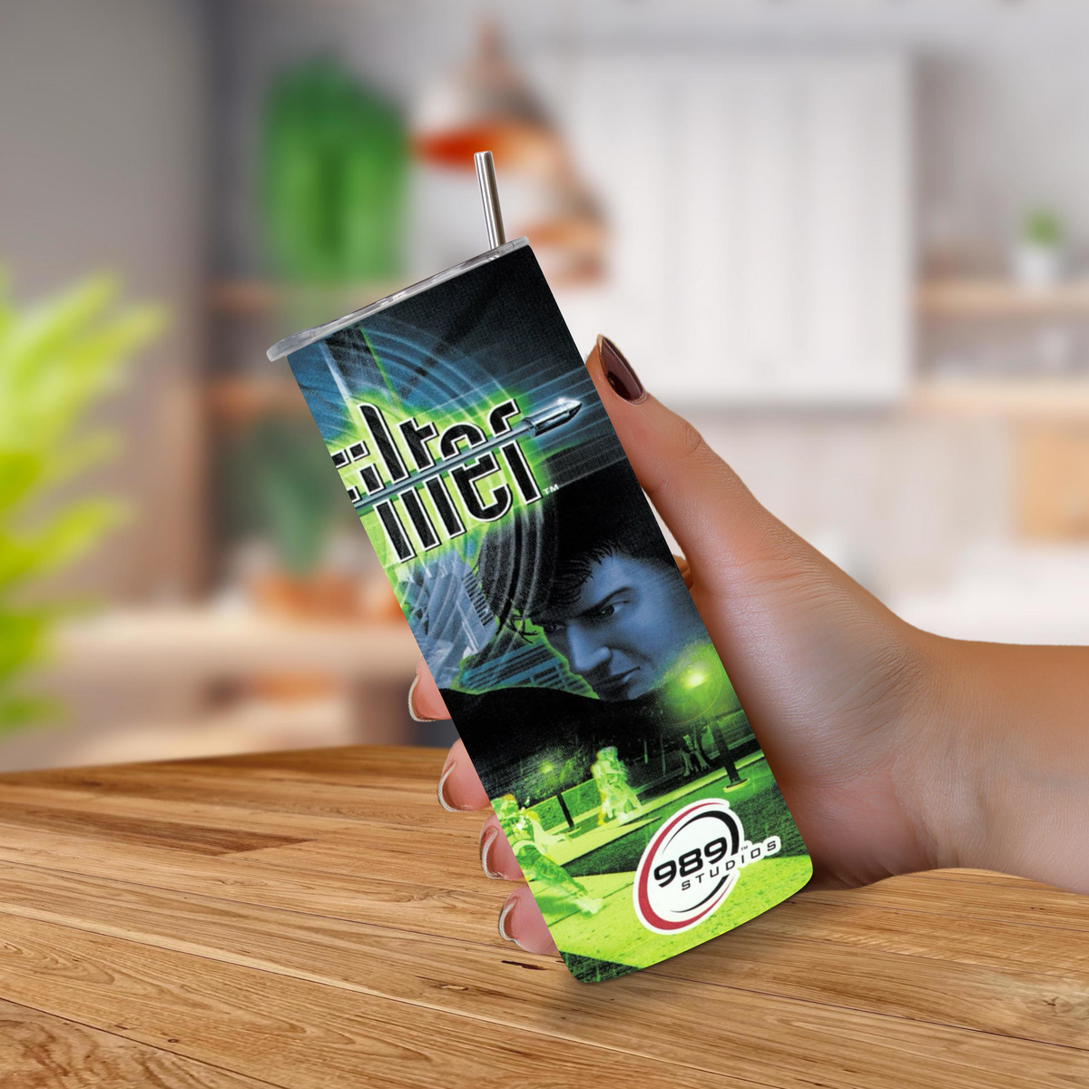 A hand holding a slim stainless-steel tumbler with a metal straw, featuring a custom Syphon Filter design & PS1 Design. The background shows a blurred indoor setting with a wooden table.