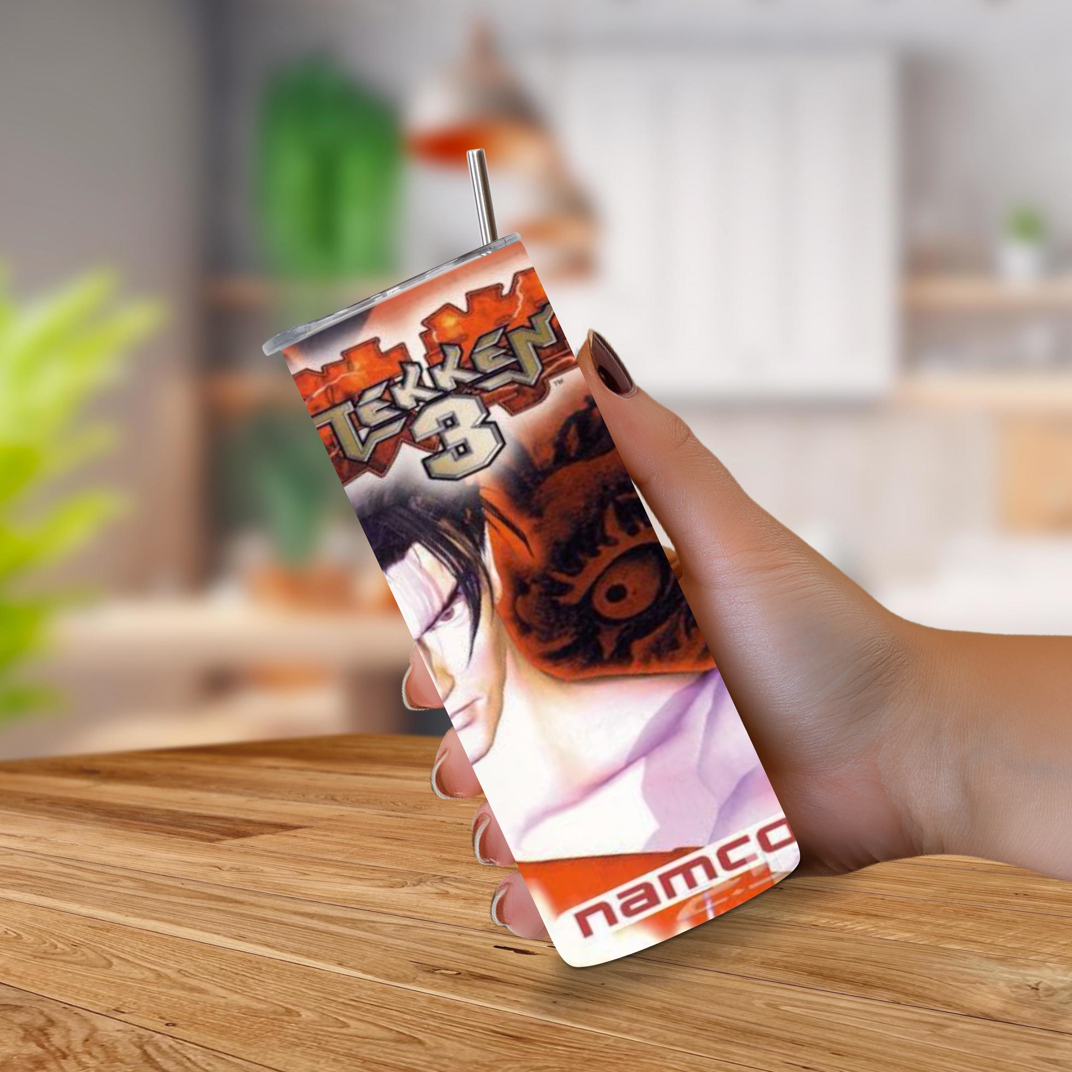A hand holding a 20oz tumbler featuring PlayStation artwork Tekken 3 with a metal straw, set against a blurred indoor background and wooden tabletop.