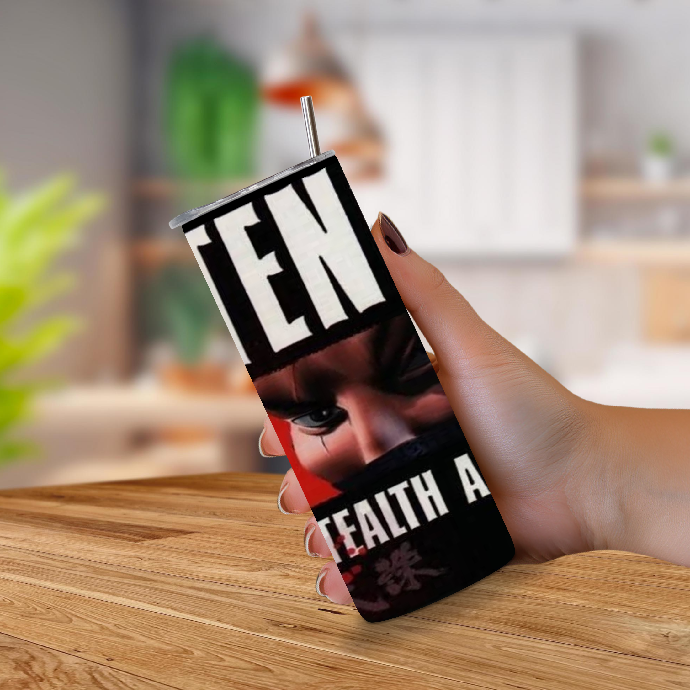 A hand holding a black 20oz insulated tumbler with a straw, featuring a graphic print of a face with a tear and bold white text reading 'TENCHU Stealth Assassins on a blurred kitchen background.