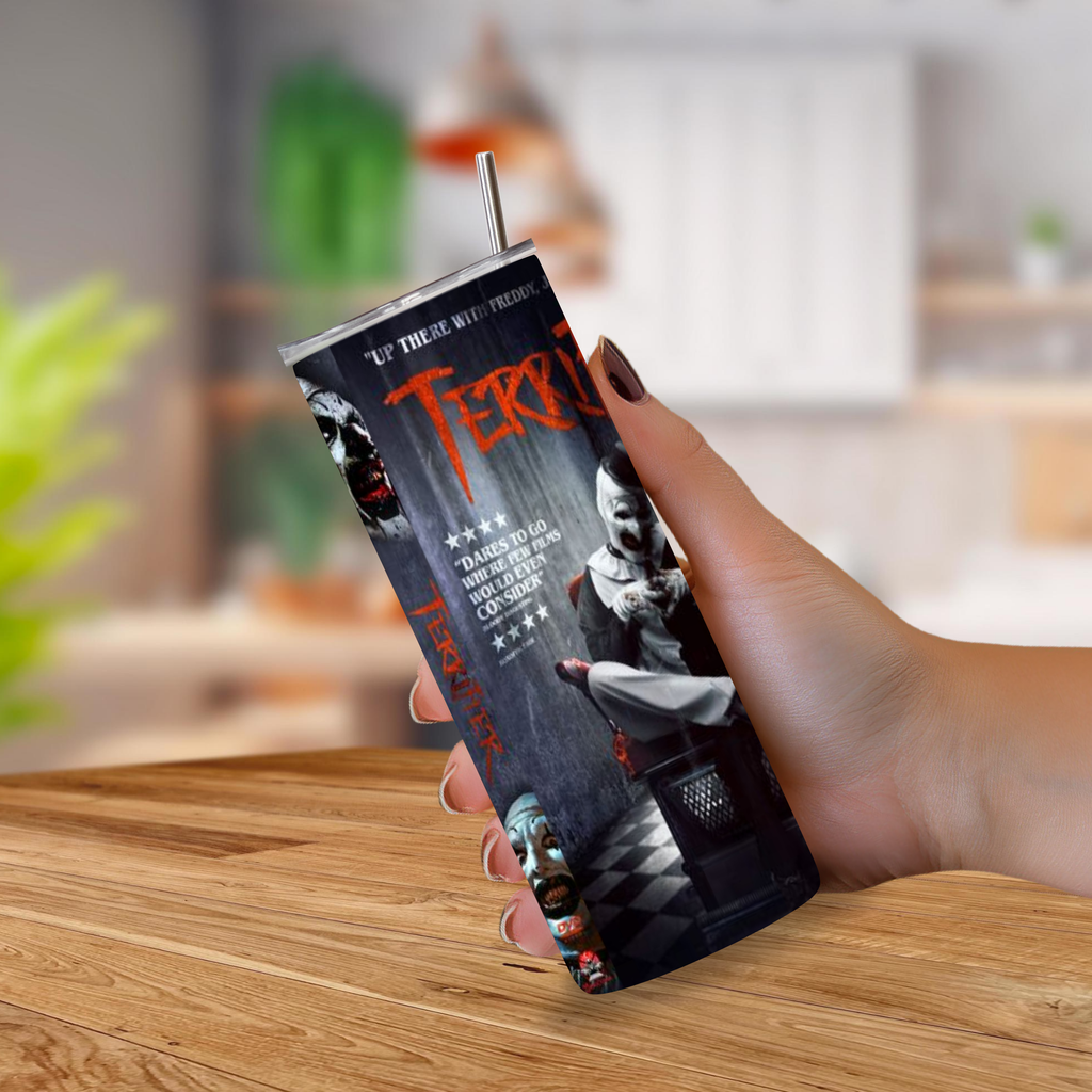 A hand holding a slim stainless-steel tumbler with a printed horror movie poster design featuring the title 'TERRIFIER' and a metal straw, set against a blurred kitchen background.