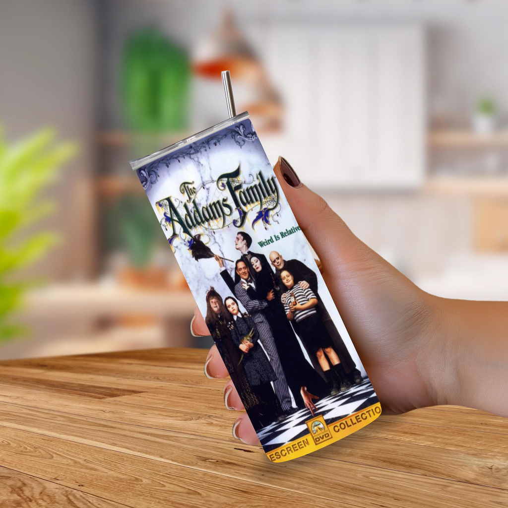 A hand holding a slim rectangular collectible tumbler featuring a printed 'The Addams Family' movie design, with a metal straw, against a blurred indoor background and wooden tabletop.