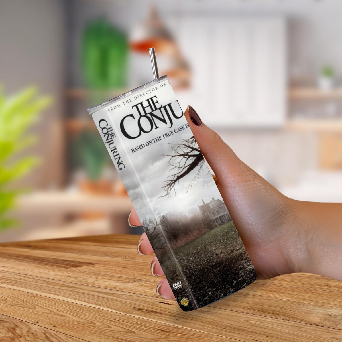 A hand holding a 20oz tumbler with a printed 'The Conjuring' movie sleeve design and a metal straw, set against a blurred kitchen background.
