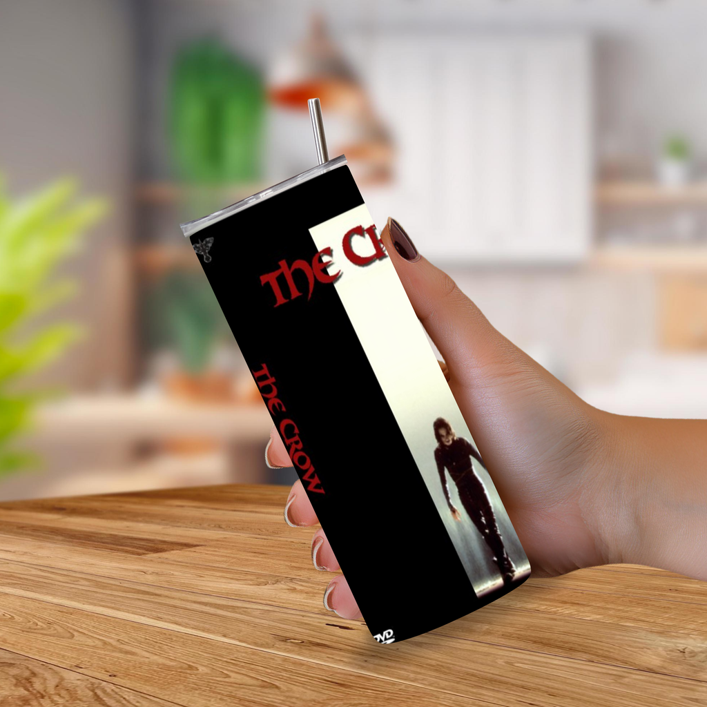 A hand holding a slim rectangular tumbler with a straw, printed with 'The Crow' artwork in black, red, and cream, set against a blurred kitchen background.