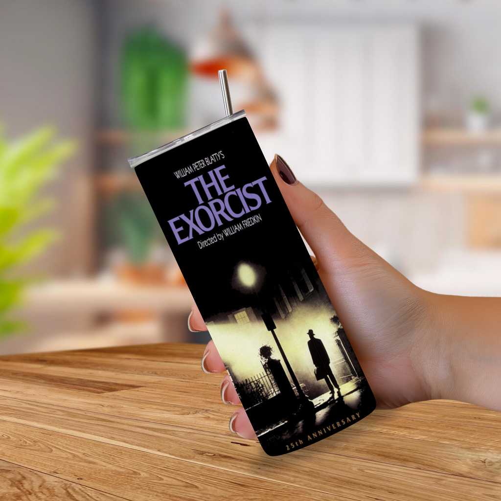 A hand holding a tall, slim tumbler with a straw featuring the 'The Exorcist' 25th Anniversary design, set against a blurred kitchen background.