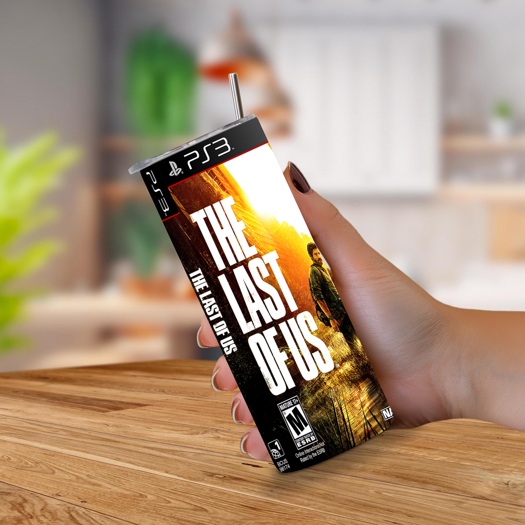 A hand holding a PS3 game cover designed to look like a 20oz stainless steel tumbler, featuring the title 'The Last of Us' and artwork from the game, set against a blurred kitchen background.