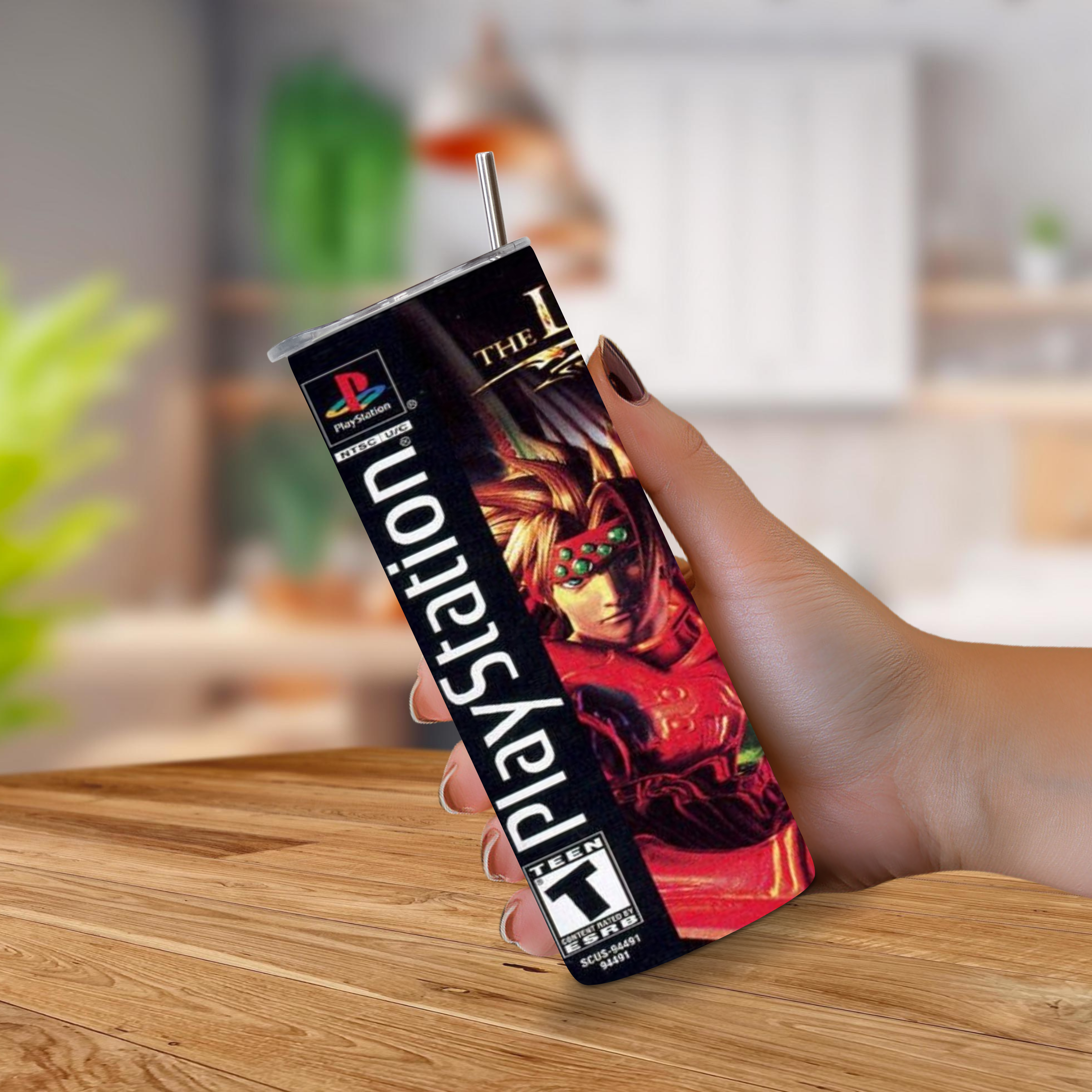 A hand holding a PlayStation-branded tumbler cup with a straw, featuring "The Legend of Dragoon" design, set against a blurred indoor background with a wooden table.