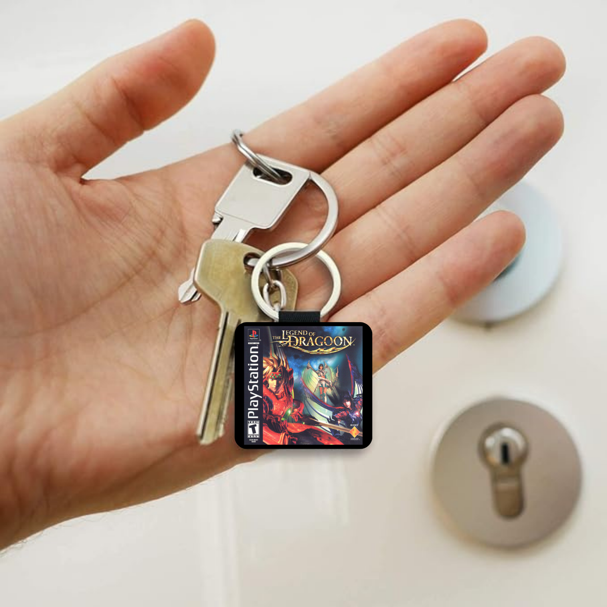 A hand holding a keychain with a square PlayStation "The Legend of Dragoon" key fob, alongside two metal keys, against a blurred background of a lock on a door.