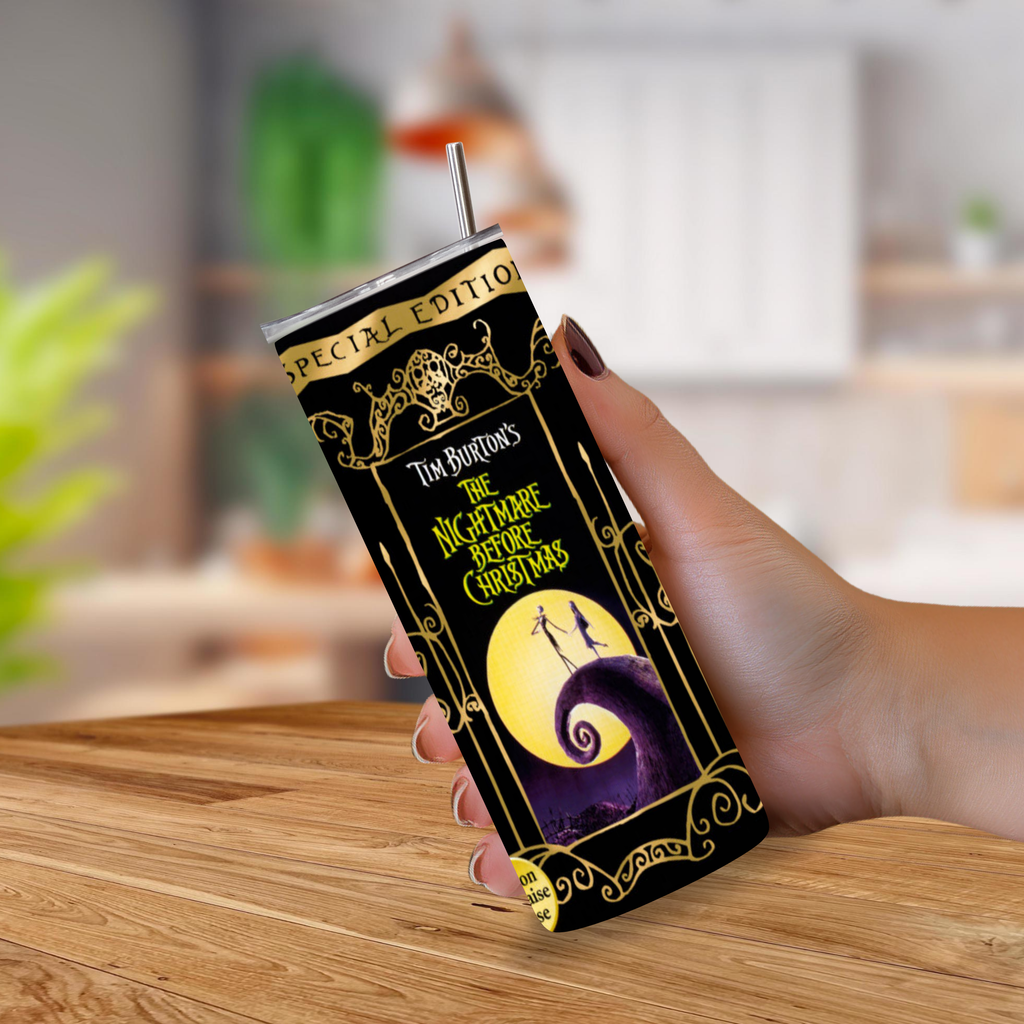 A hand holding a black insulated tumbler with a metal straw, printed with 'The Nightmare Before Christmas' artwork in gold, yellow, and purple, set against a blurred indoor background.